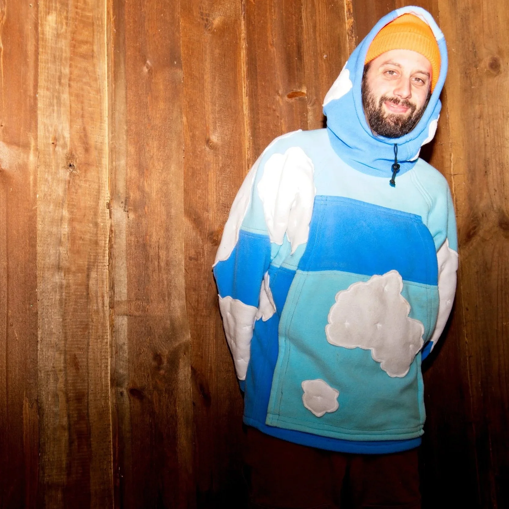 Twelve Clouds with a beard wearing a blue and white hoodie with clouds stitched on it and an orange beanie standing against a wooden wall.