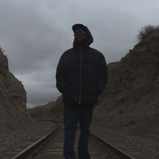 That Kid Nova standing on railroad tracks between rocky hills on a cloudy day wearing a jacket and bandana.