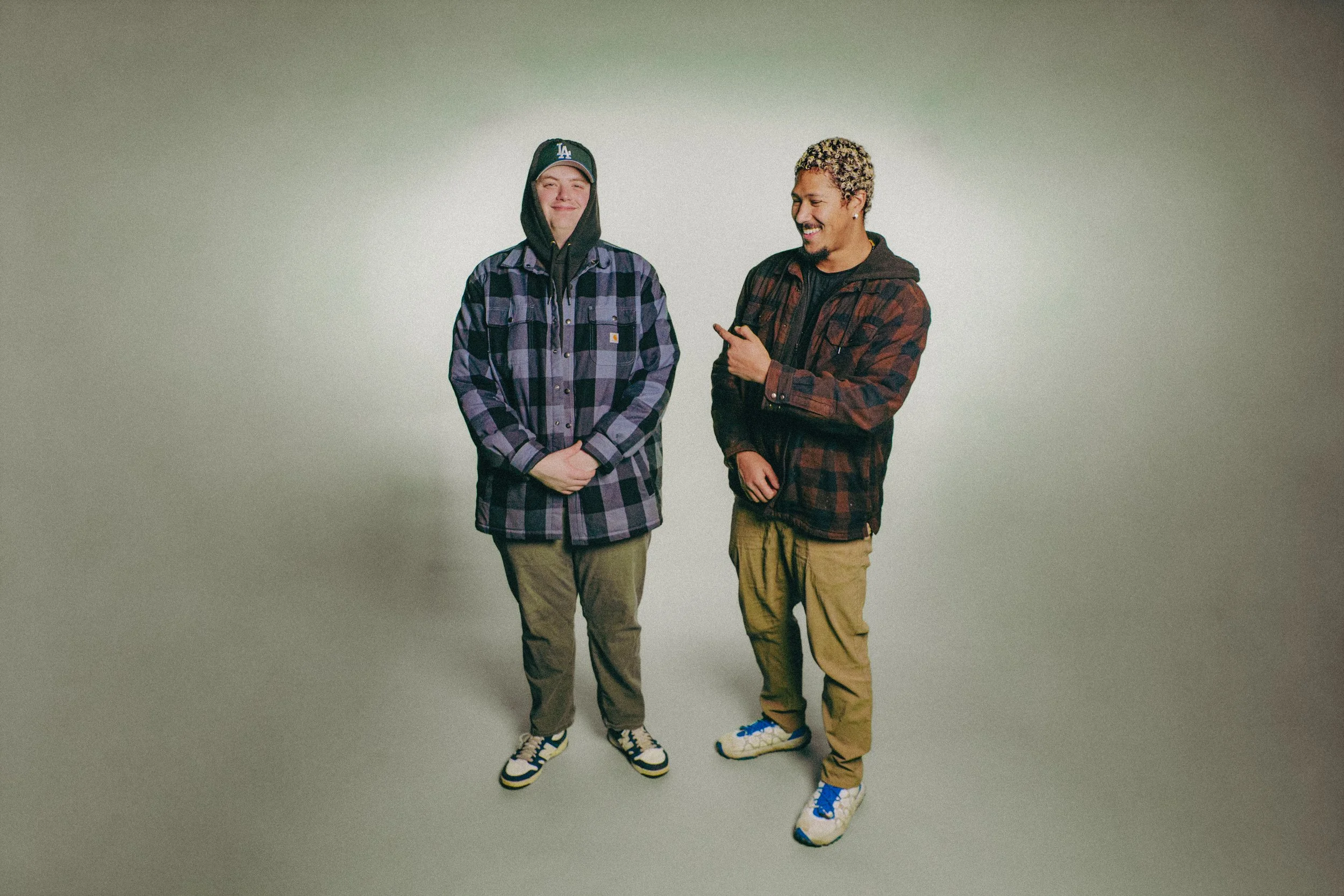 Mellow Mike and R'Know during a photoshoot.  The background is a gray gradient with Mellow on the left and R'Know on the right.  Both are wearing checkered jackets and Mellow has his hood up with a baseball cap.  R'Know is smiling and pointing.