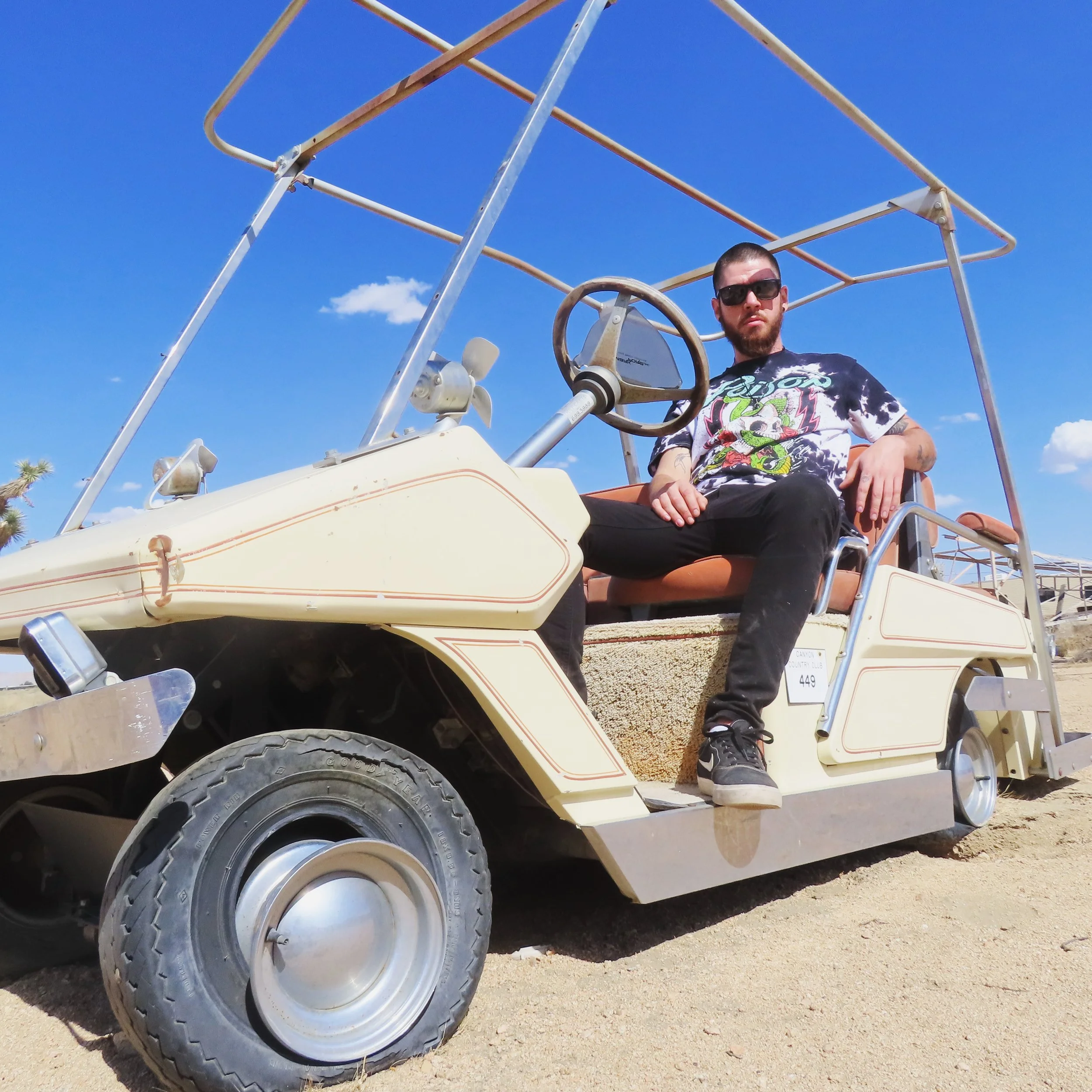 Yung Signal with tattoos and sunglasses sitting in a golf cart with a metal frame, parked on a dirt ground under a bright blue sky with some clouds.