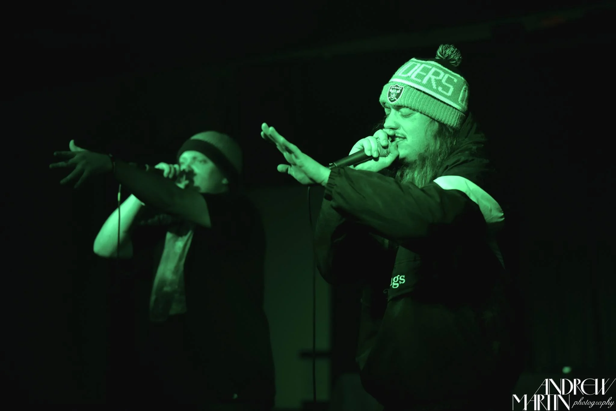 Symon Palmer and Petey Peak performing on stage together.  Both have mics in their hands with a green stage light shining on them.