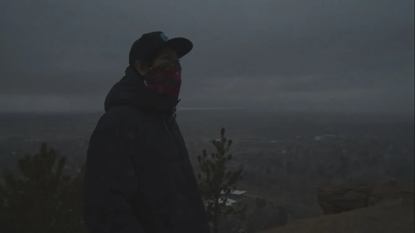 That Kid Nova wearing dark jacket, cap, and bandana standing outdoors on a cloudy day, overlooking the Billings, MT cityscape from the rims.