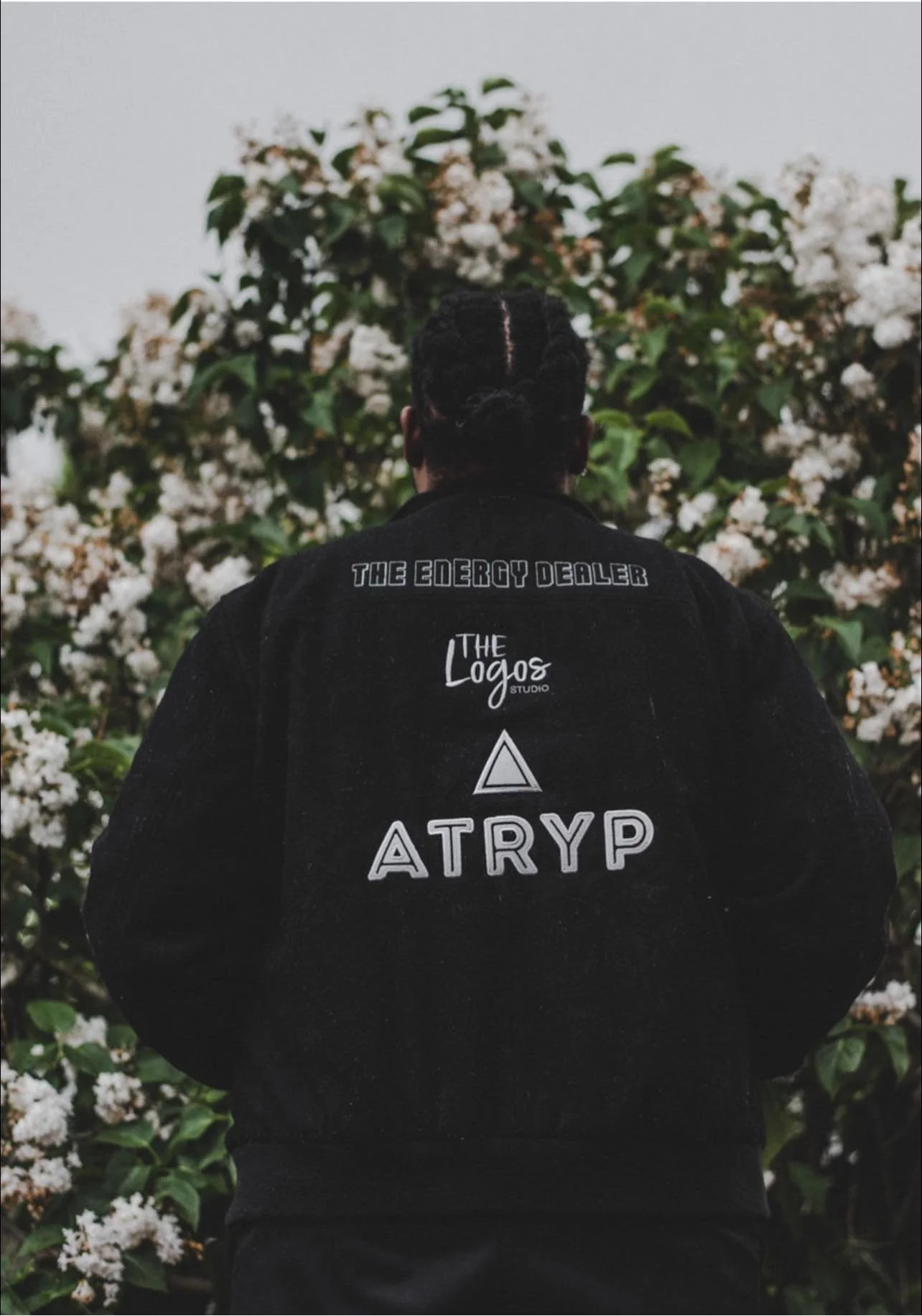 Back view of a person with braided hair wearing a black jacket with white text on the back, standing in front of a bush with white flowers.