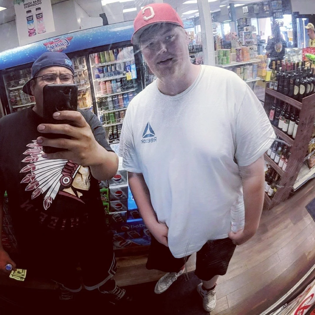 The man on the right is wearing a white t-shirt and a red cap, while the man on the left is wearing glasses and a navy cap, holding the phone. Behind them are shelves with drinks, including sodas and bottles of alcohol.