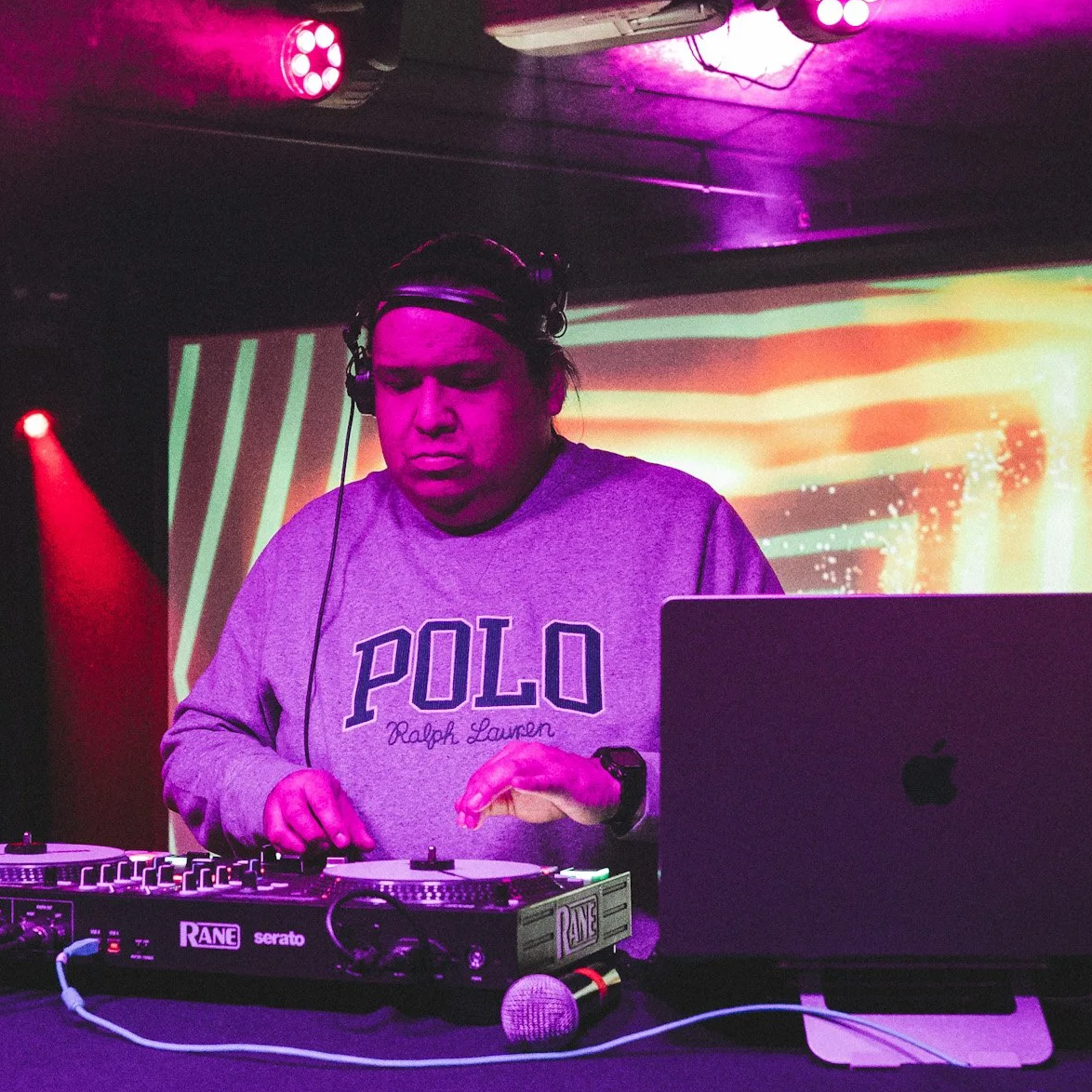 Dean Smif (aka DJ Exodus) performing, wearing headphones and a gray POLO Ralph Lauren sweatshirt, in a dimly lit club with colorful pink, purple, and orange lighting and a digital backdrop.