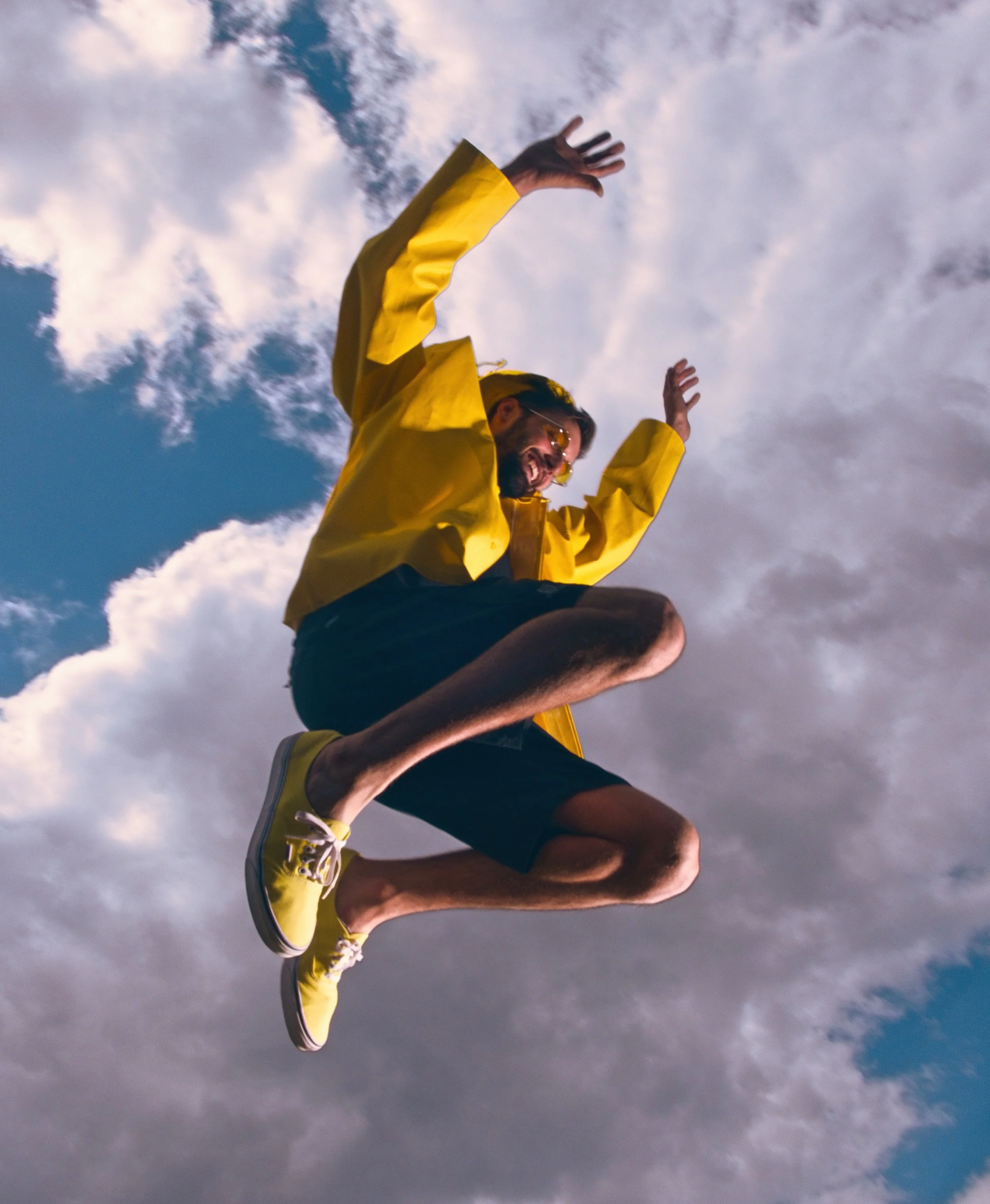 Twelve Clouds in a yellow raincoat and yellow shoes with white soles jumping or falling in the sky with clouds and blue sky behind him.