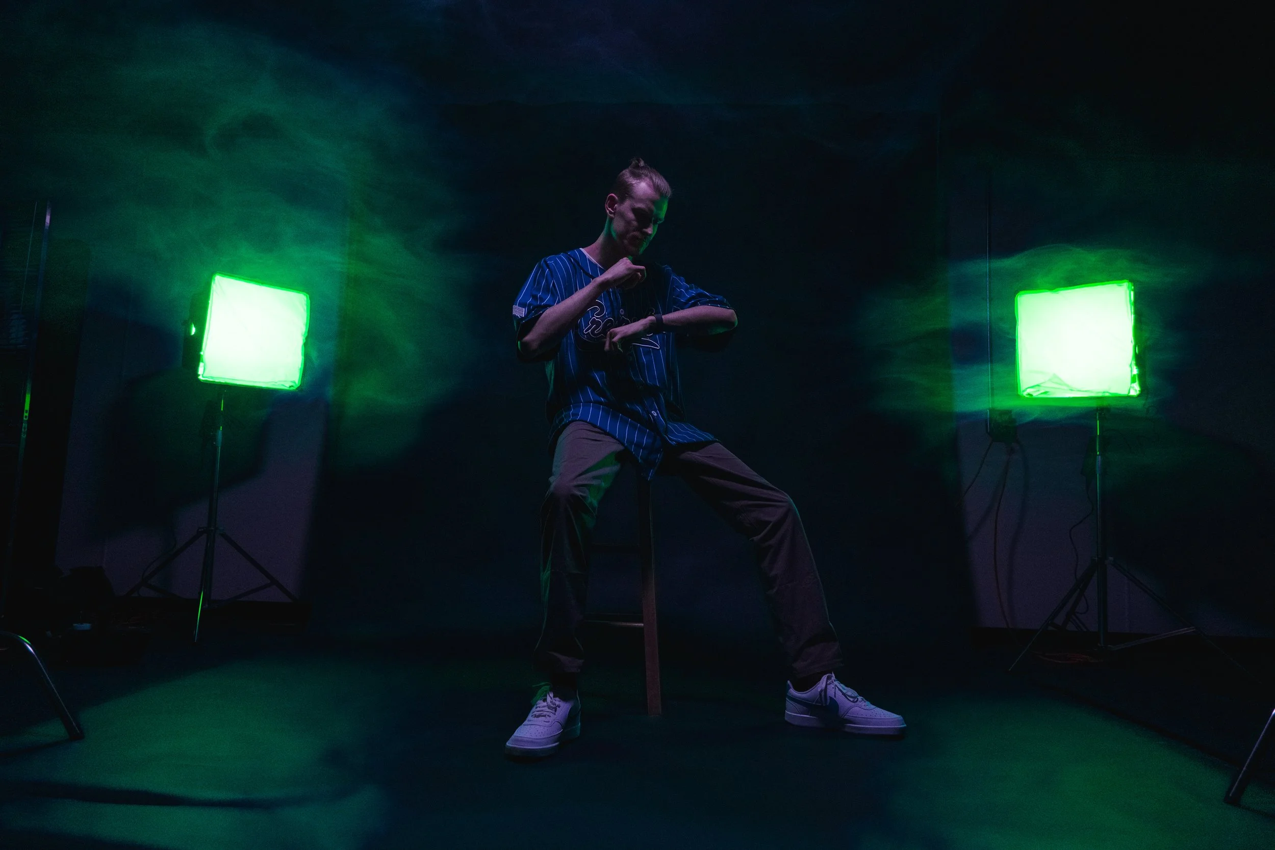 TR. in a baseball jersey and tan pants pose in a dark room with green lighting and green LED lights.