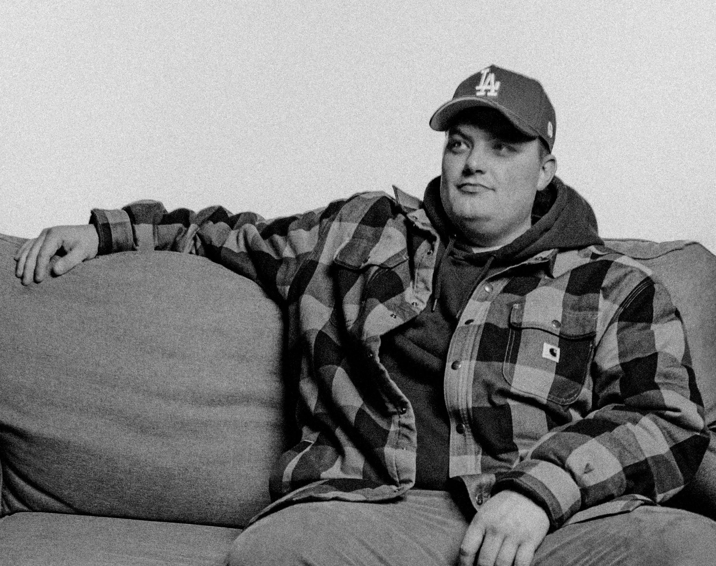 Mellow Mike sitting on a couch, wearing a baseball cap and a checkered jacket, looking to the side.