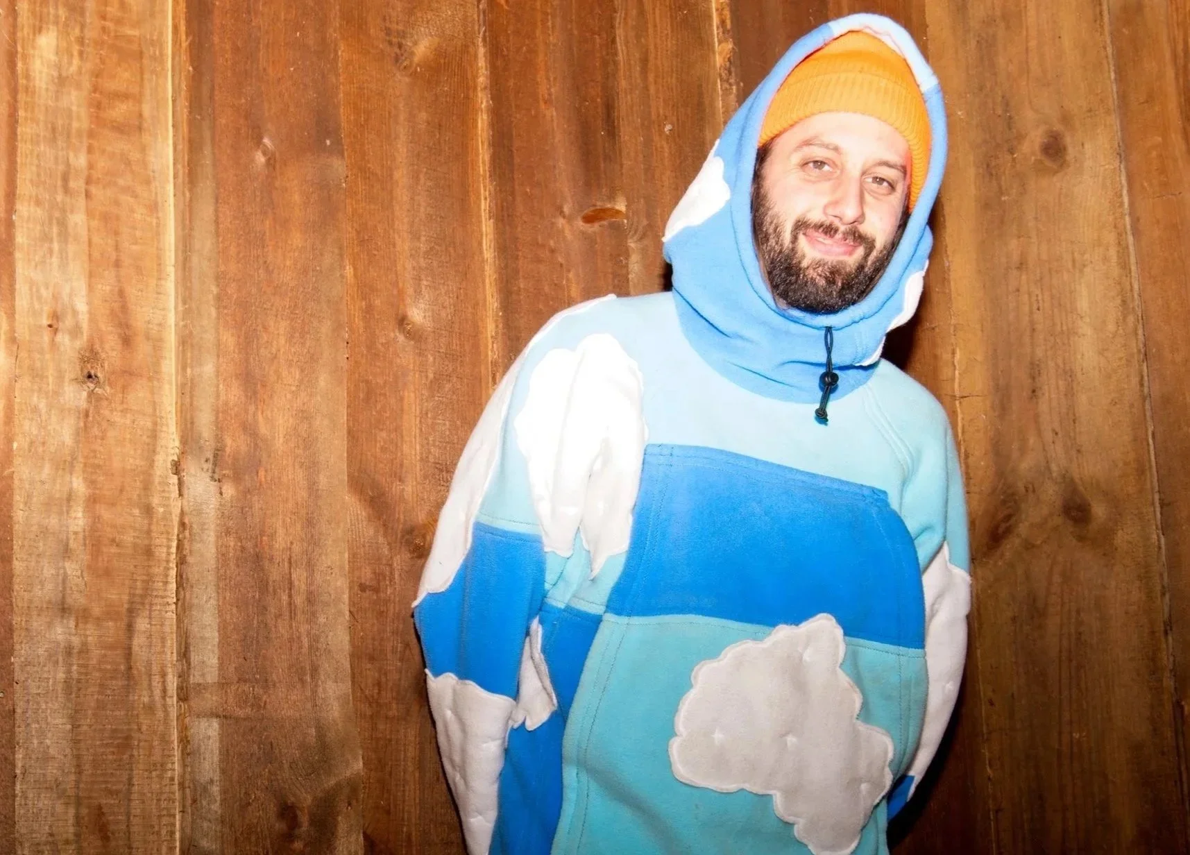 Twelve Clouds with a beard wearing a blue and white cloud-themed hoodie and an orange beanie, standing in front of a wooden wall.