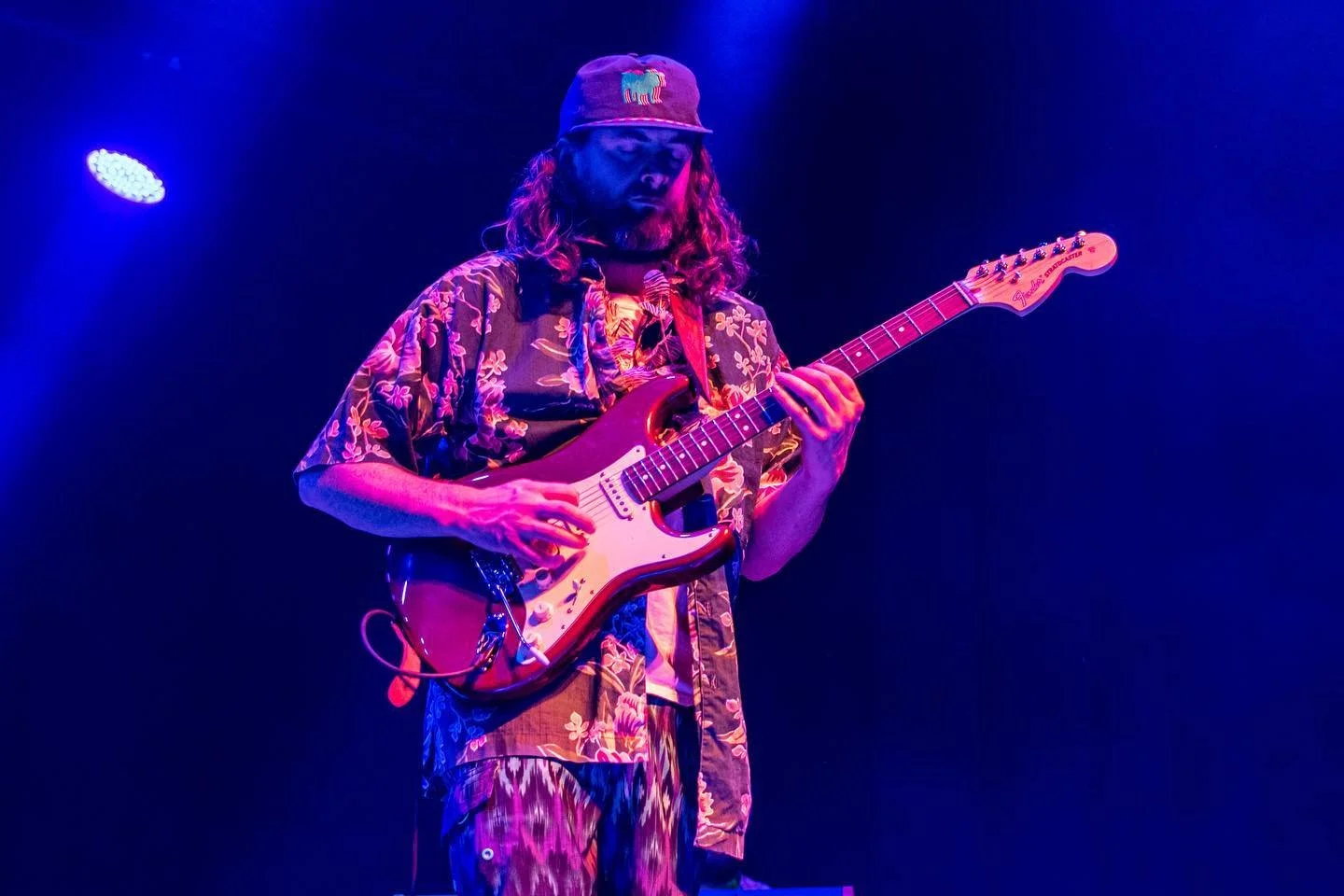 Chris from STiLGONE playing an electric guitar on stage under purple and blue lighting, wearing a floral shirt, patterned pants, and a baseball cap with a colorful logo.