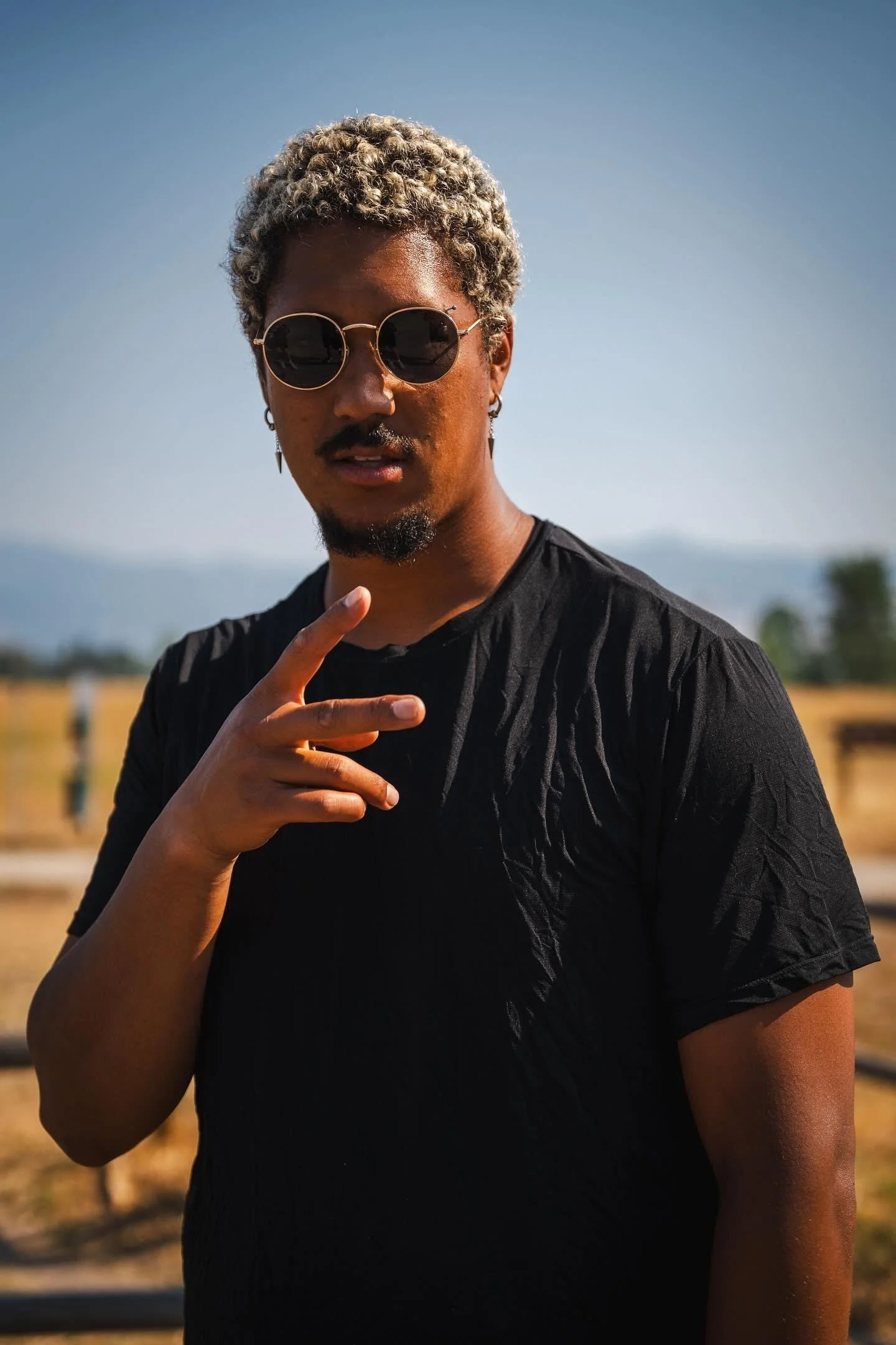 Photo of R'Know, wearing round sunglasses and a black shirt, standing outdoors during daytime, with a clear blue sky in the background.