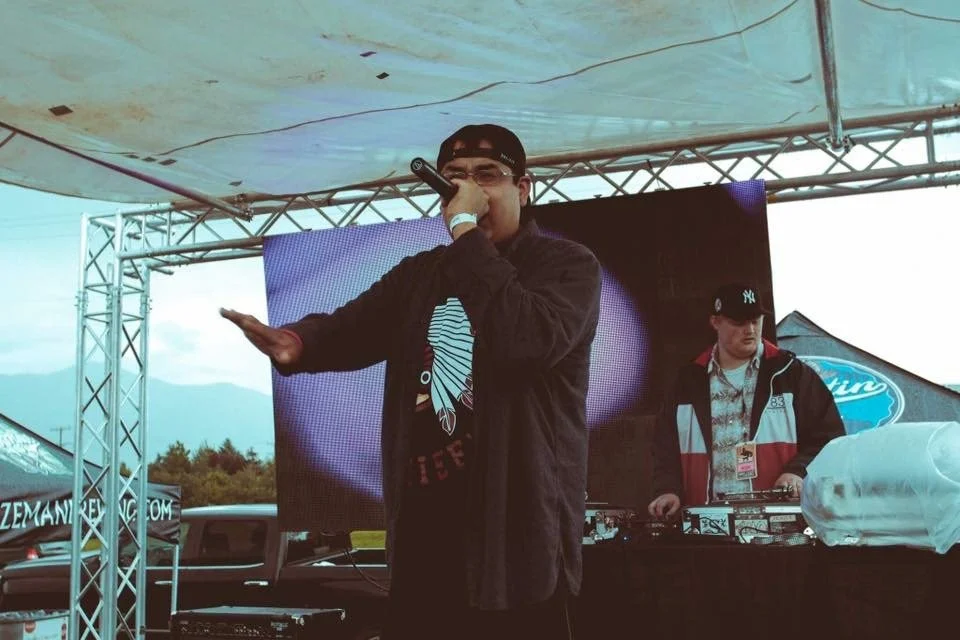 C. Boy and Mellow performing at a block party in Bozeman.  C. Boy is rapping with a mic in his hand and Mellow is behind the turntables, with a screen and mountain range in the background.