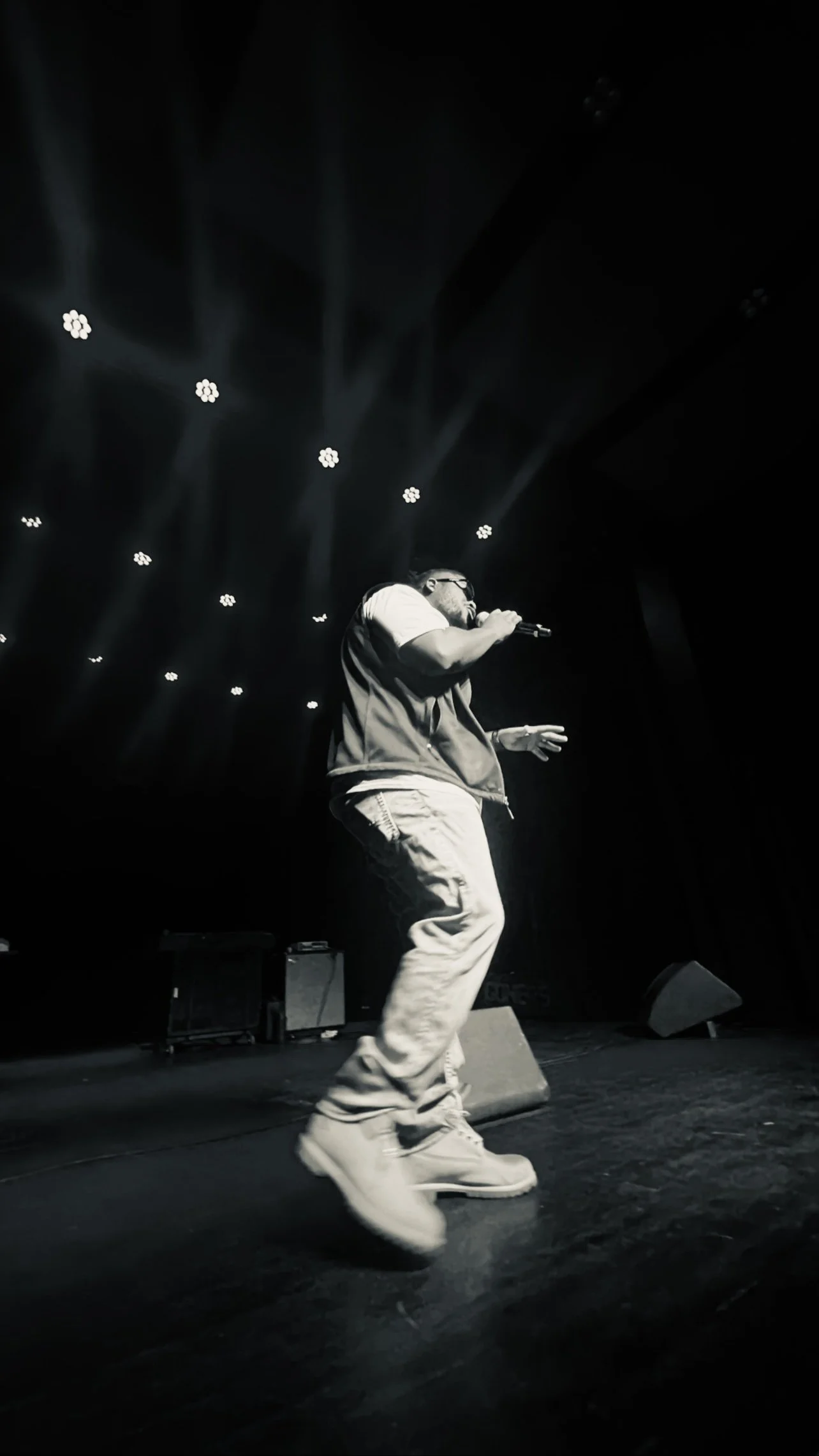 Atryp performing on stage, dressed in a vest, baggy pants, and boots, holding a microphone with stage lights overhead.