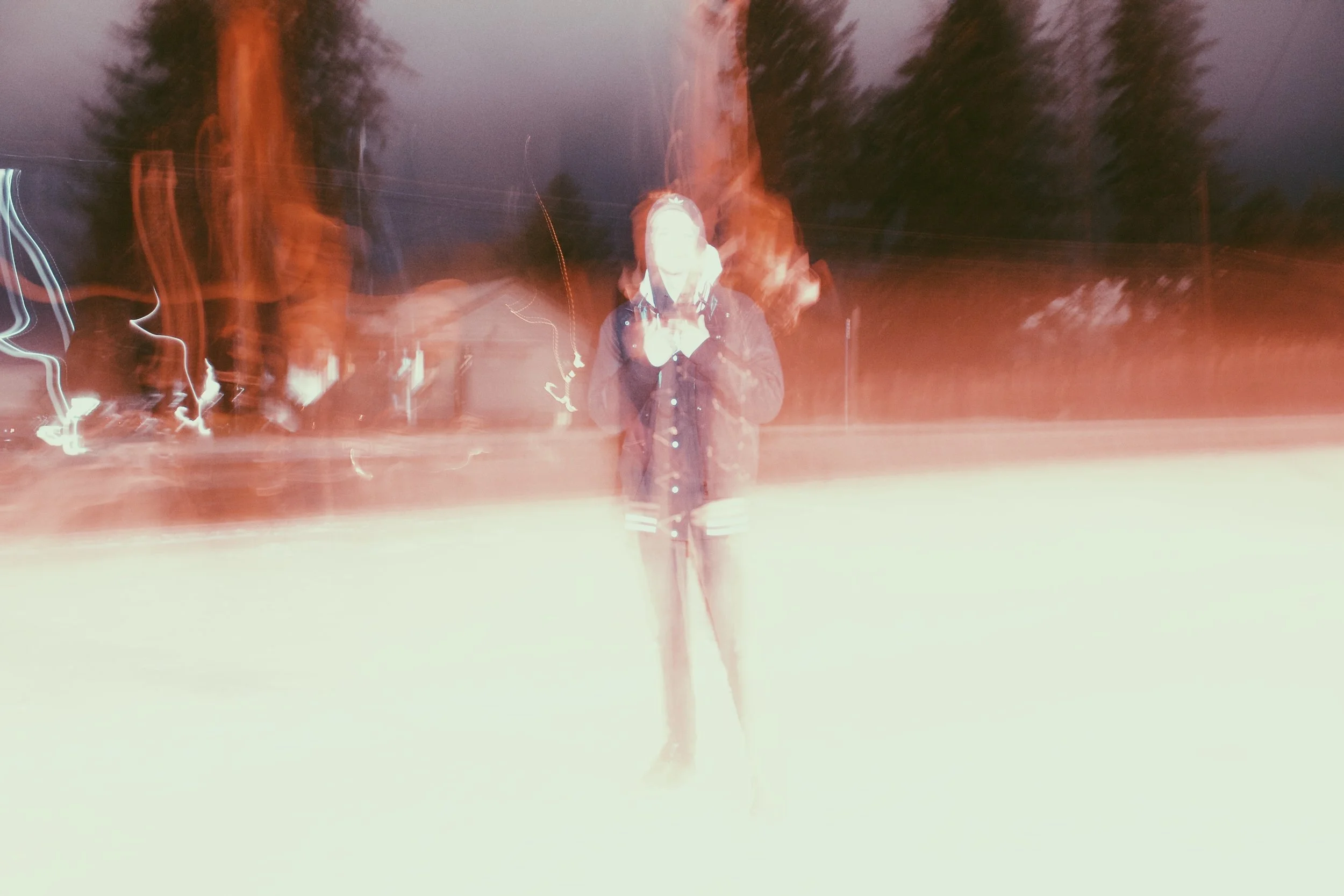 TR. in dark clothing standing outdoors at night, with motion blur and light trails surrounding them.