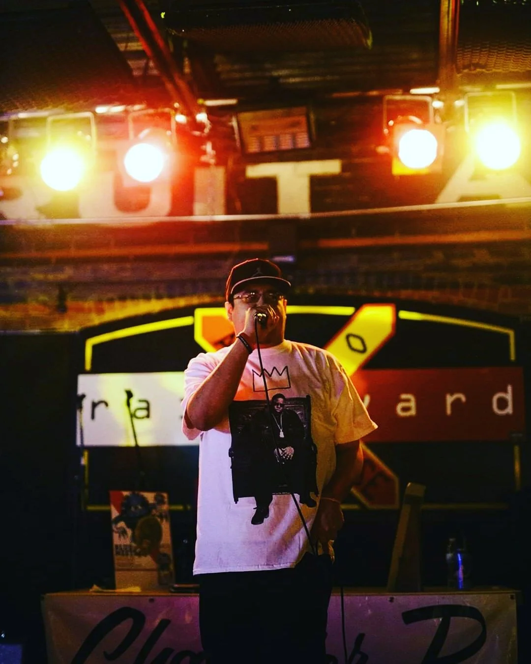 C. Boy rapping on stage with bright lights behind him, wearing a white T-shirt featuring a graphic print and a dark cap, in a dimly lit bar or club setting.