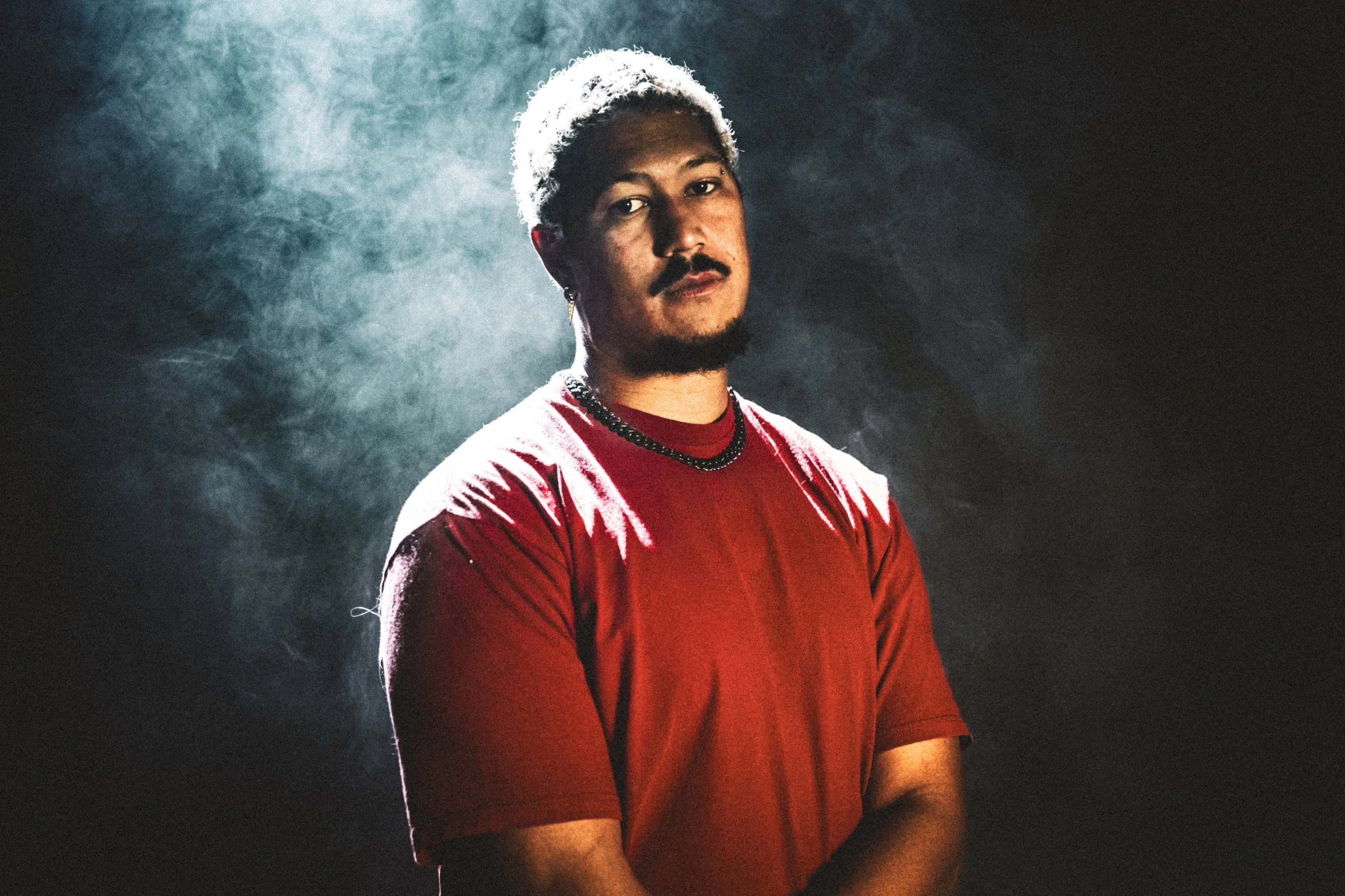 R'Know wearing a red shirt, chain necklace, and earrings, with a mustache and beard, standing against a dark background with moody lighting and presence of smoke or mist.