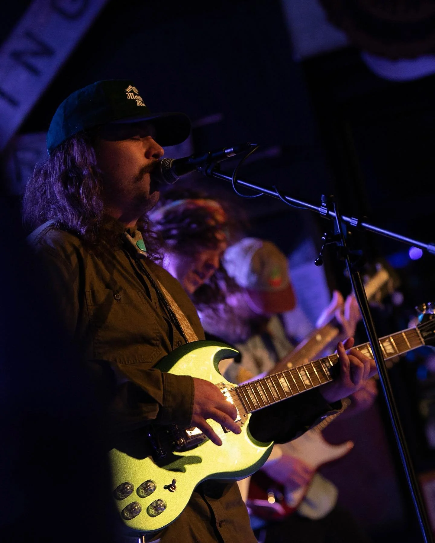 STiLGONE frontman Symon Palmer, wearing a green cap, singing into a microphone while playing a yellow electric guitar in a dark, atmospheric setting with purple and blue lighting.