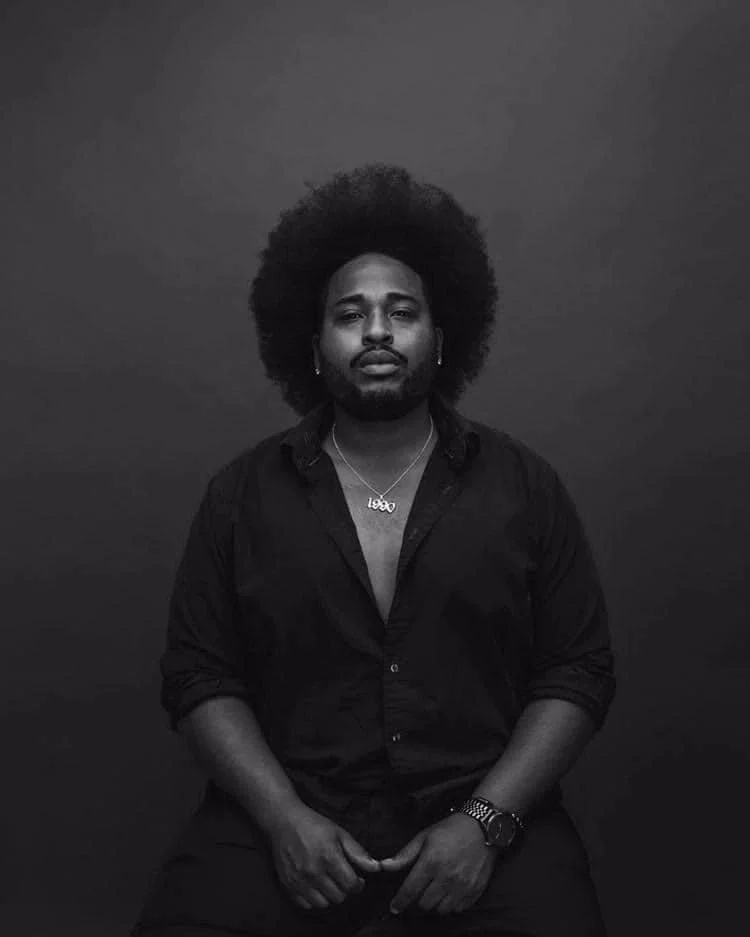 Black and white photo of Atryp with an afro hairstyle, wearing a black shirt with the top buttons open, a necklace, and a wristwatch, sitting against a plain dark background.