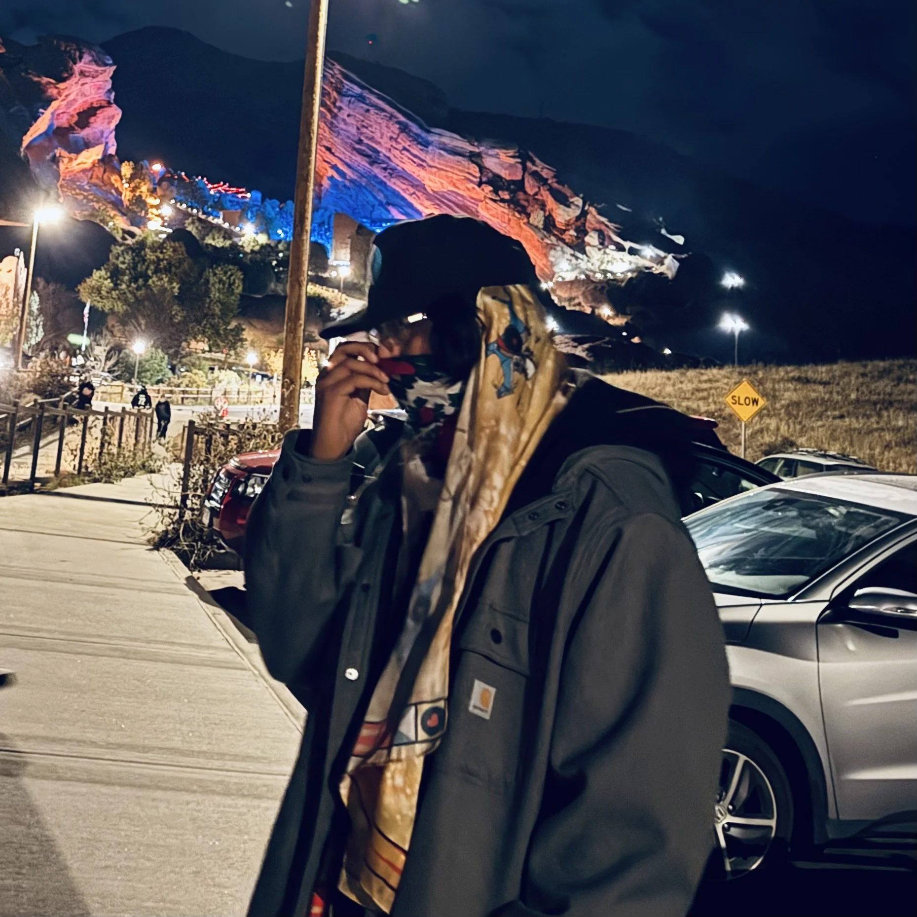 That Kid Nova, standing sideways wearing a jacket, long scarf, baseball hat, and glasses.  He's adjusting the bandana on his face, pulling it up with the Red Rocks Amphitheater in the background.