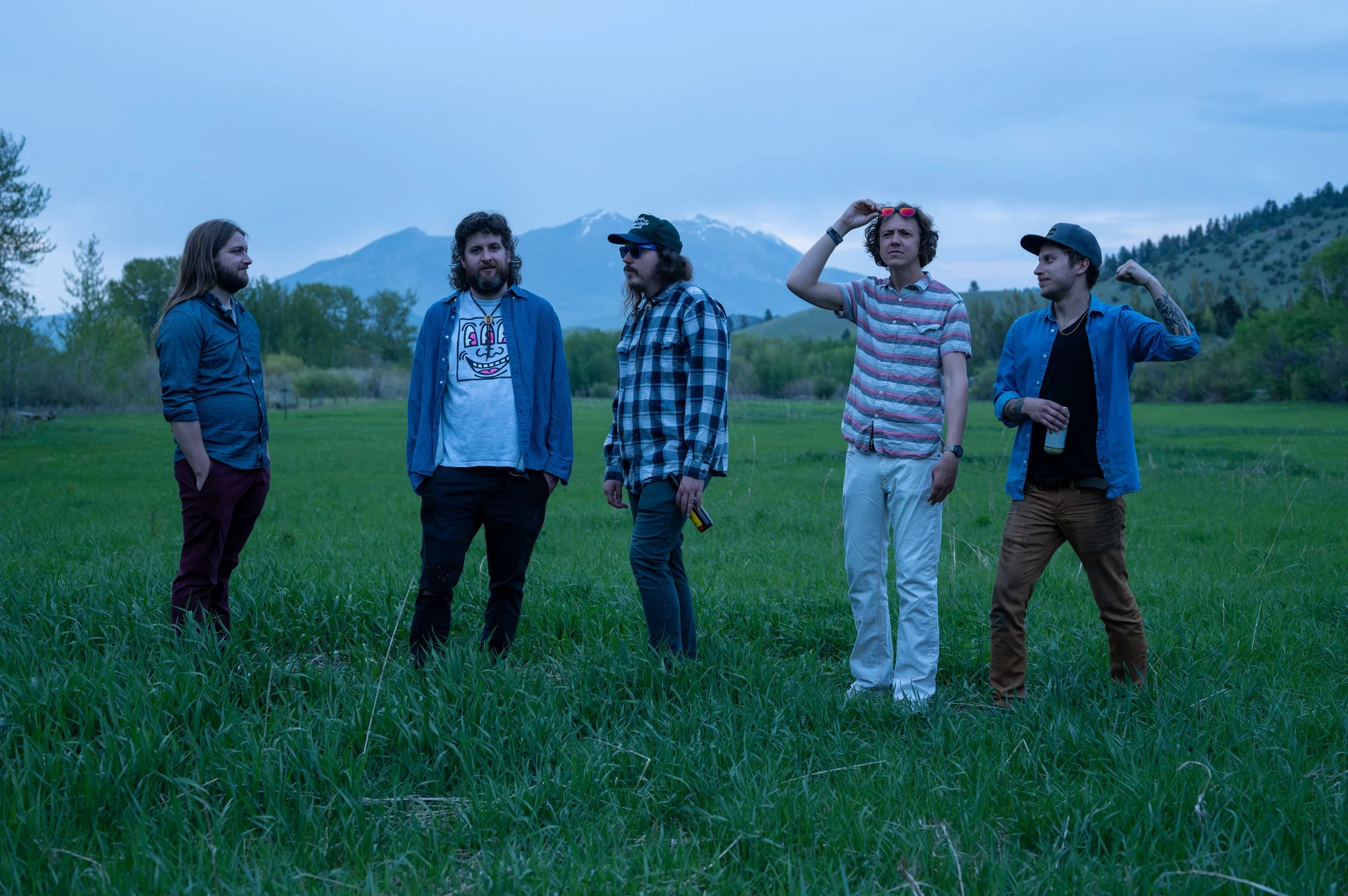 Five men stand in a grassy field with mountains in the background during dusk or dawn. They are dressed casually, with some wearing plaid shirts and sunglasses. One of them is adjusting his sunglasses while others are engaging in conversation or looking around.