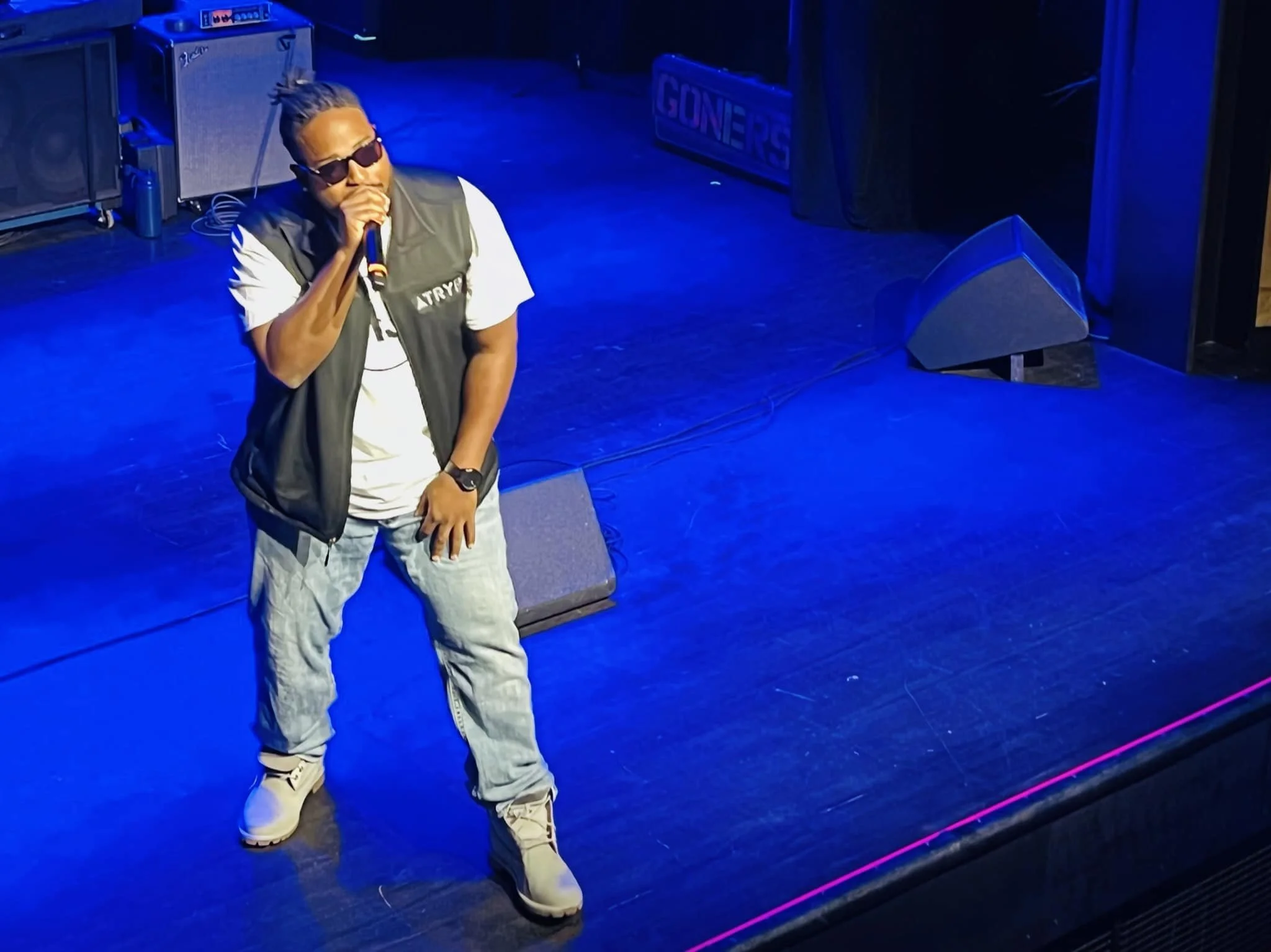 Atryp performing with sunglasses holding a microphone on a blue-lit stage, wearing a white T-shirt, grey vest, and jeans.
