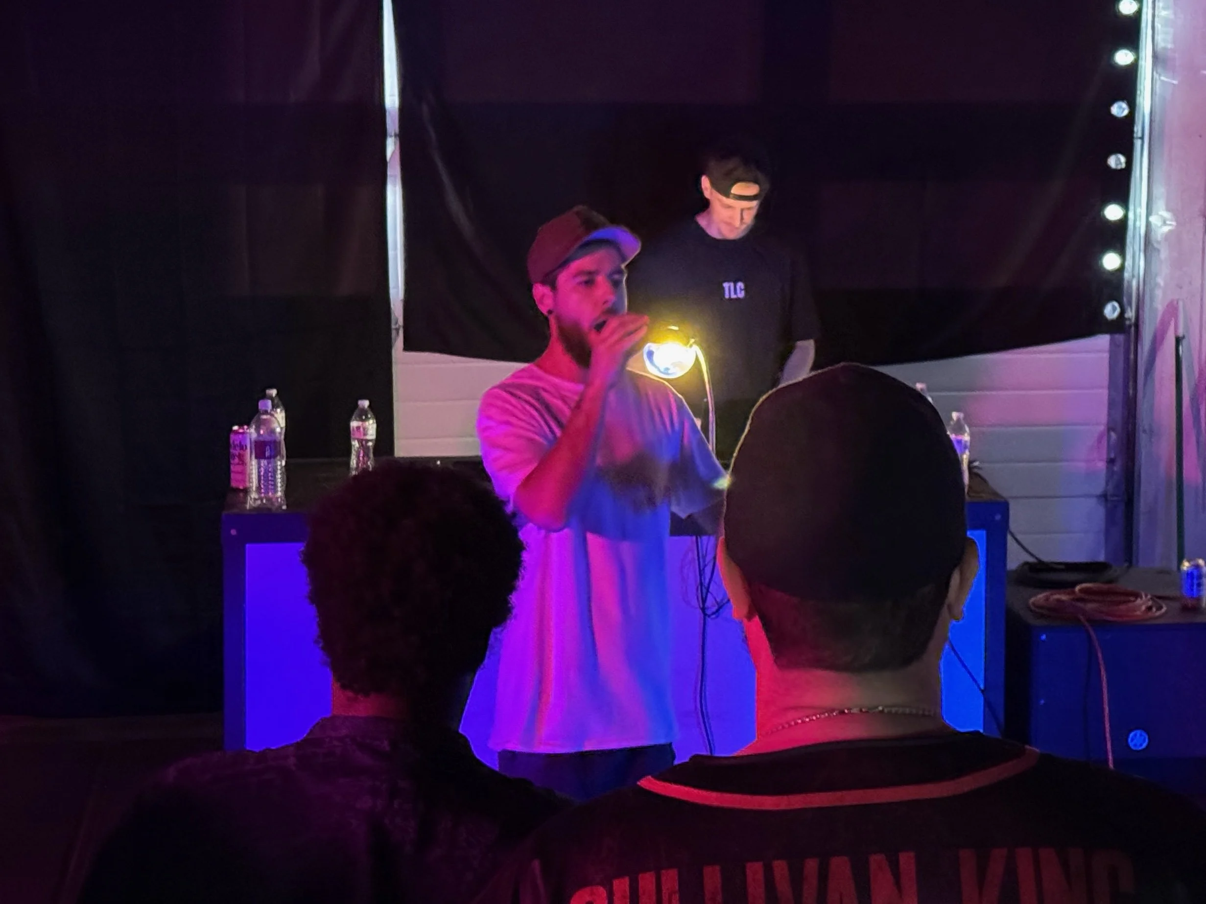 Two young men standing in front of a black backdrop, illuminated by purple, pink, and yellow stage lighting, with tables and bottles of water in the foreground, performing at a nighttime event or party.