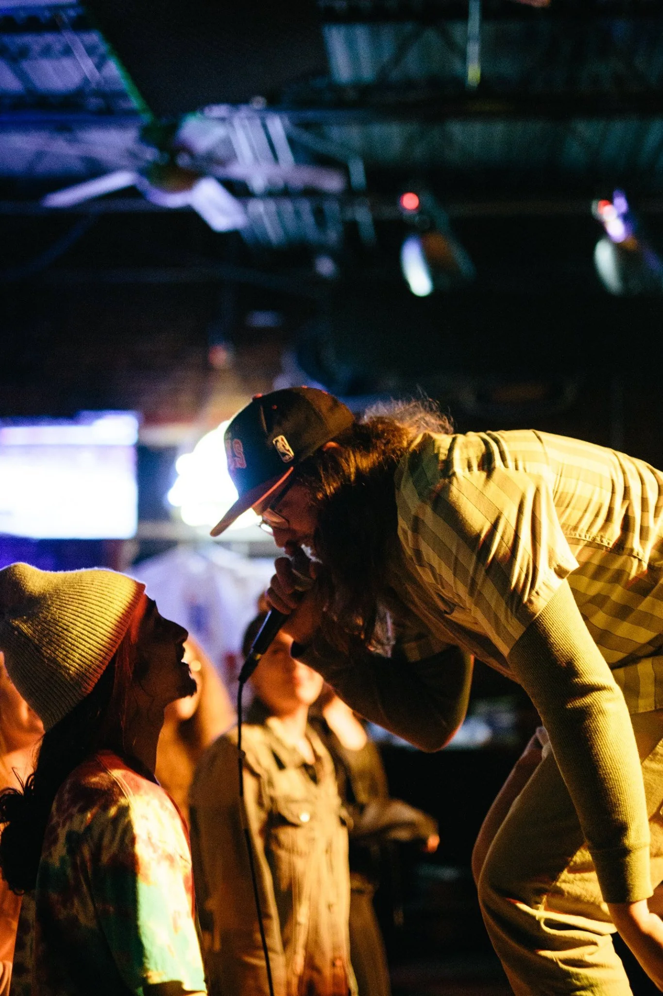 Symon Palmer wearing a baseball cap, glasses, and a button up shirt performs while up close, face to face with a fan in the crowd.