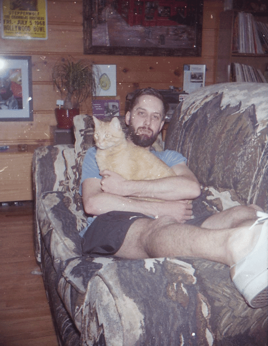 Twelve Clouds sitting on a patterned couch holding an orange tabby cat, in a wood-paneled room decorated with framed photographs, a potted plant, and various books.