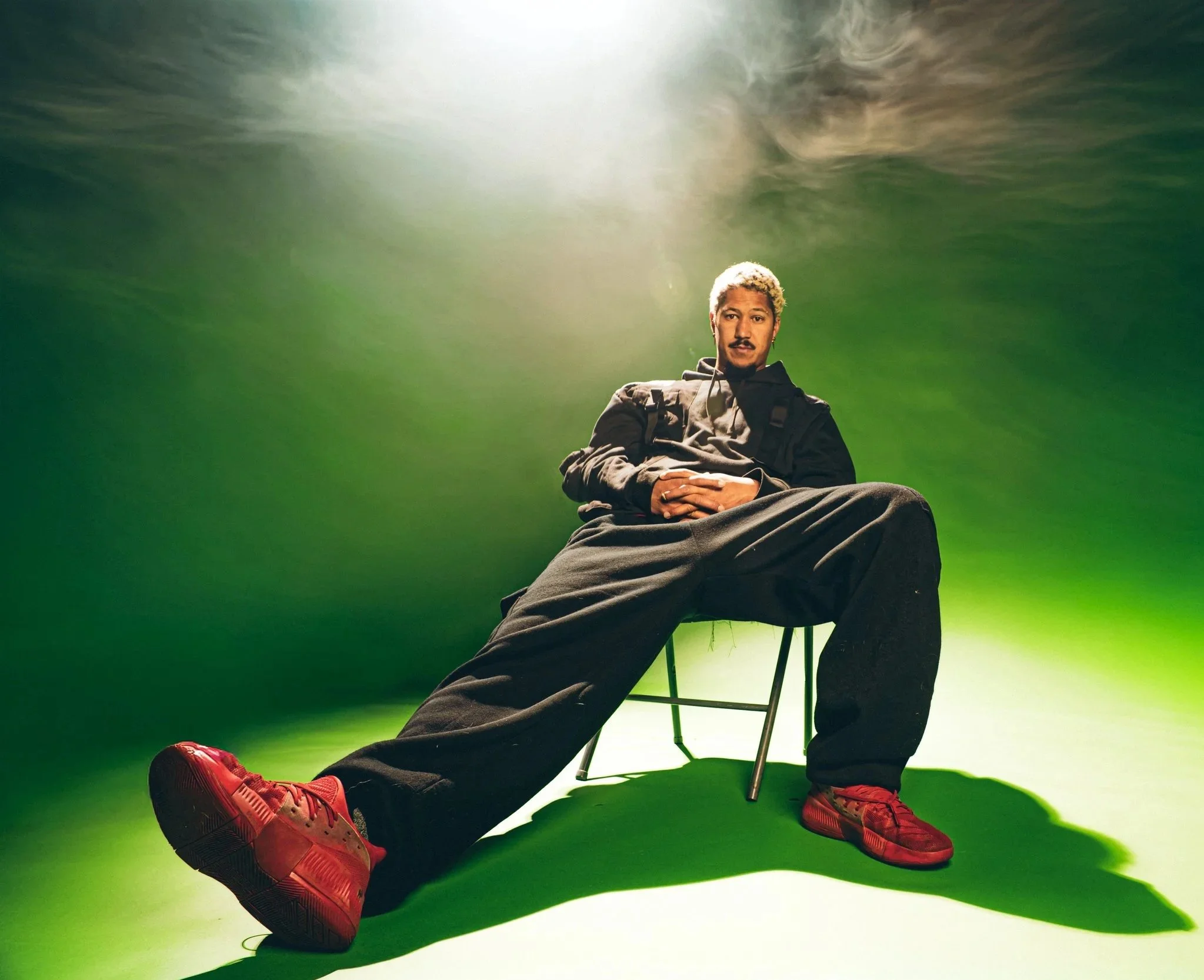 R'Know with blond hair and a mustache sitting on a black chair against a green screen background. He is wearing a dark hoodie, grey pants, and red sneakers. The lighting is bright from above, creating a dramatic effect.