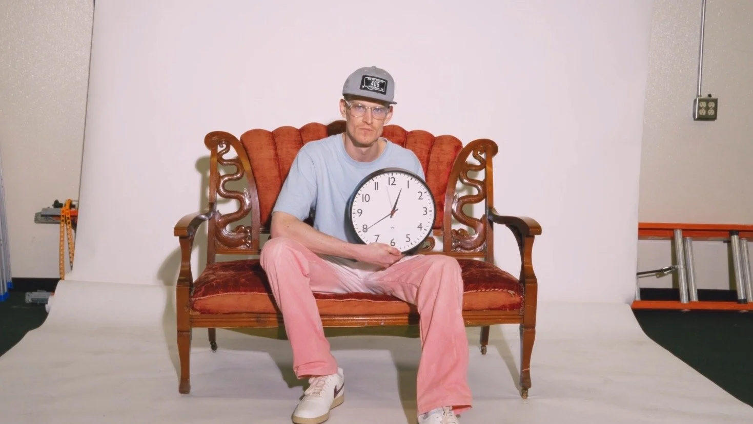 TR sitting on an ornate wooden bench holding a large round clock. He wears a gray baseball cap, glasses, a light blue t-shirt, pink pants, and white sneakers. The background includes a plain white backdrop, a ladder, and some equipment.