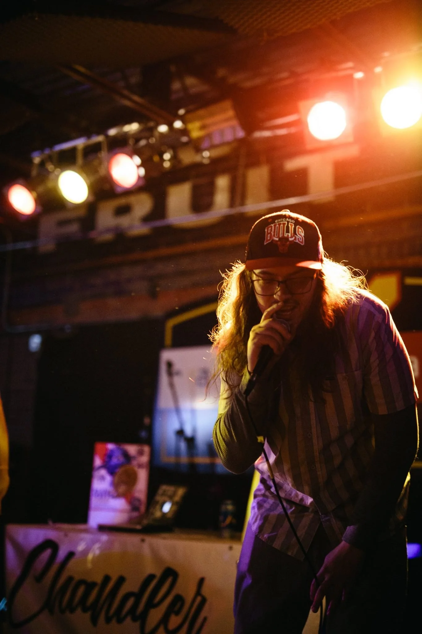 Symon Palmer with long hair wearing glasses, a Chicago Bulls cap, and a plaid shirt, holding a microphone and performing on stage with colorful lights overhead.
