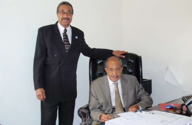 Two men, one sitting at a desk and the other standing next to him, in an office setting. The man standing is wearing a black suit, white shirt, and patterned tie, with his hand resting on the back of the seated man's chair. The seated man is wearing a gray suit, white shirt, and light-colored tie, and is holding glasses in his hand.