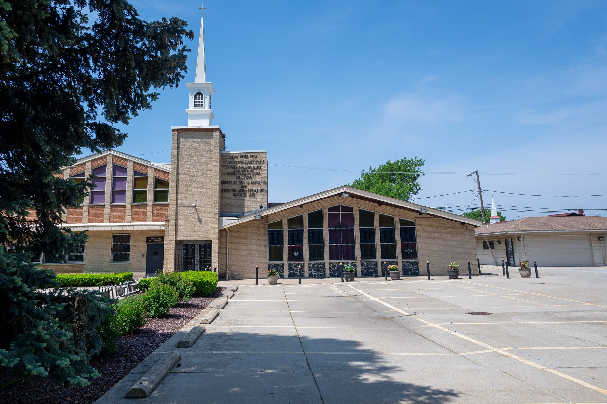 A church with a tall white steeple and a brick facade, adjacent to a parking lot with marked spaces and potted plants, surrounded by trees and overhead power lines, under a blue sky.