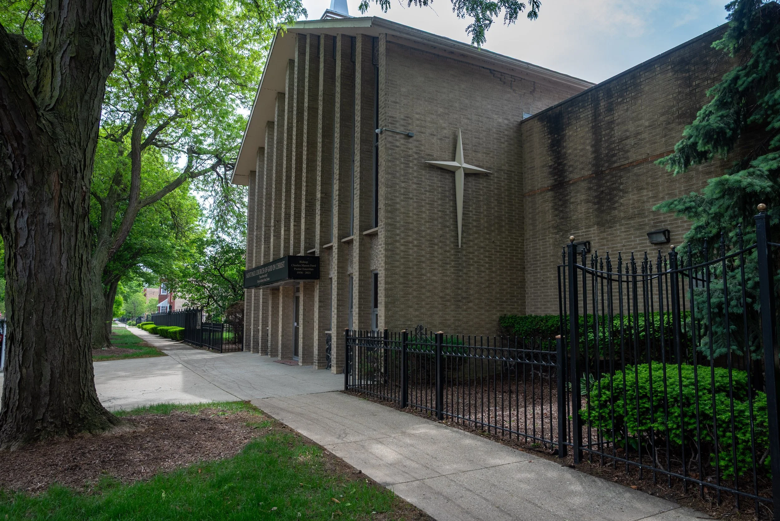 Exterior view of a church with a large white cross mounted on a brick wall, surrounded by trees and a black metal fence, with a sidewalk and lush green grass.