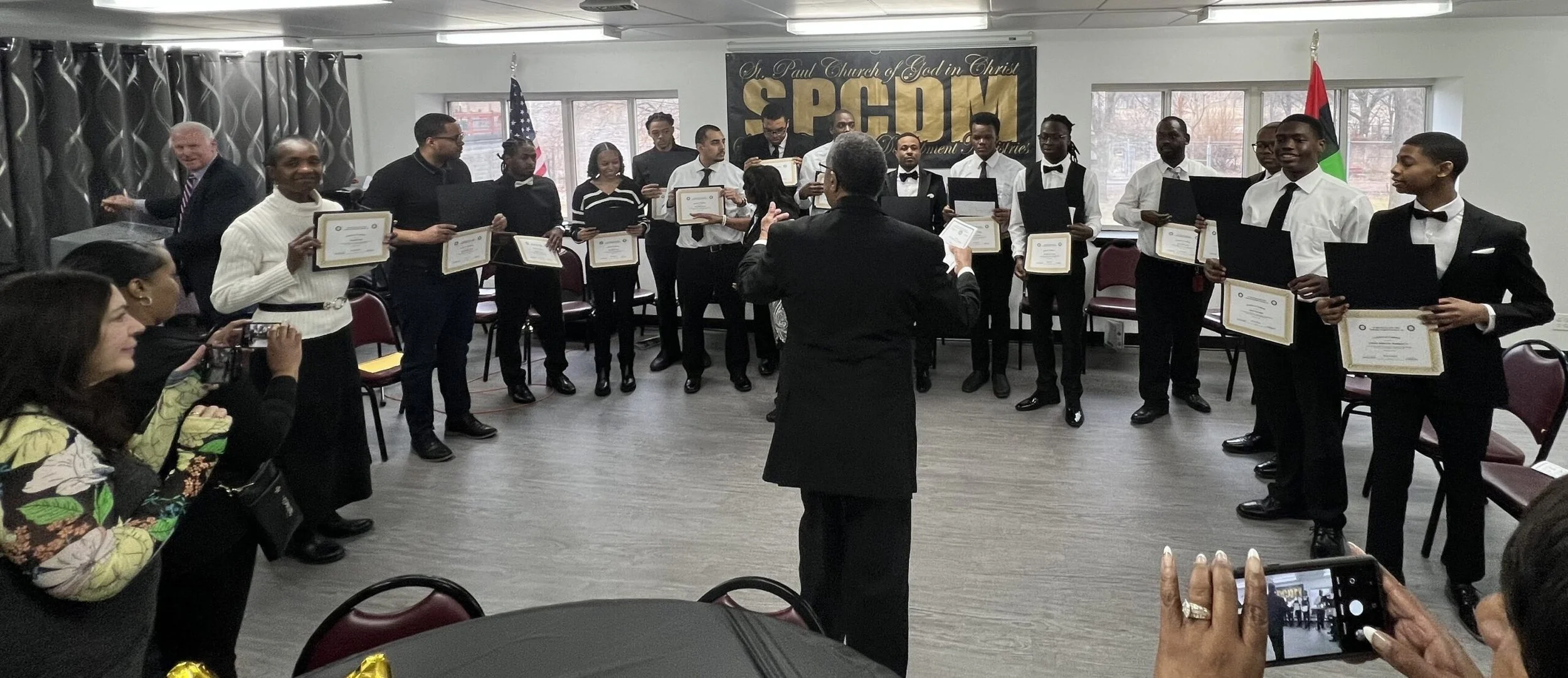 A group of young men and women dressed in formal attire standing in a semi-circle inside a room, receiving certificates from a person in front. There are chairs and a banner with the acronym 'SPCQM' in the background.