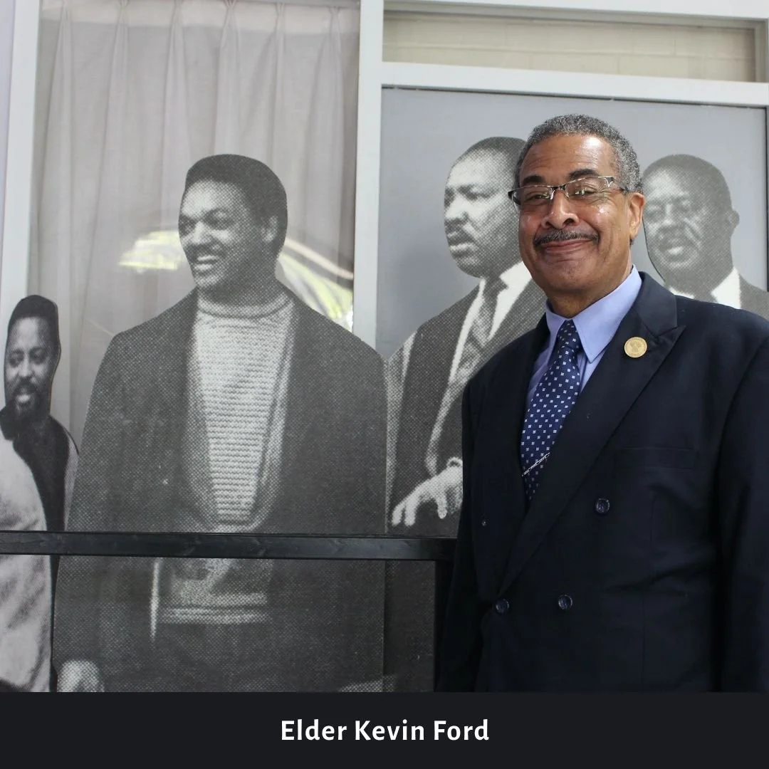 Elder Kevin Ford standing in front of a large black and white photograph of four people, smiling and dressed in formal attire, during a public or professional event.
