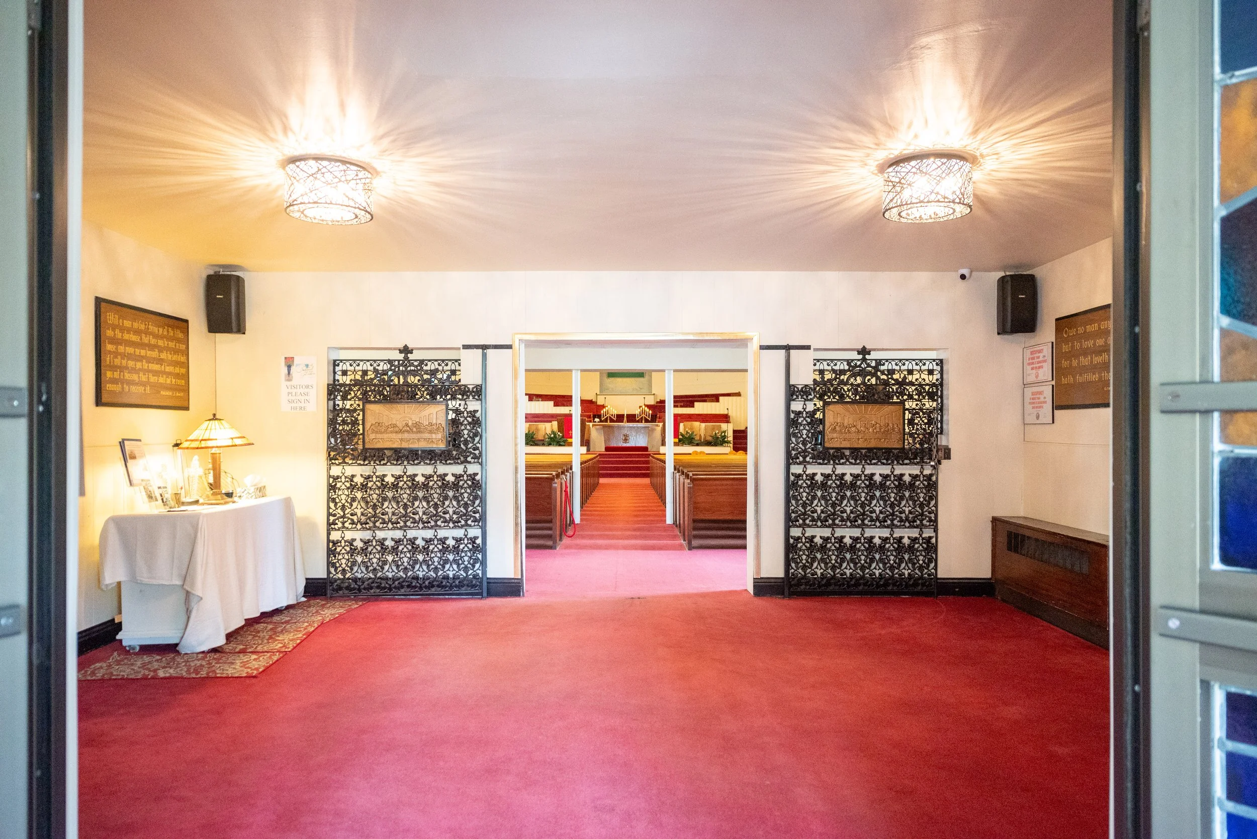 View of the entrance to a church or chapel with red carpet leading to the altar, ornate black metal gates, and wooden pews inside.