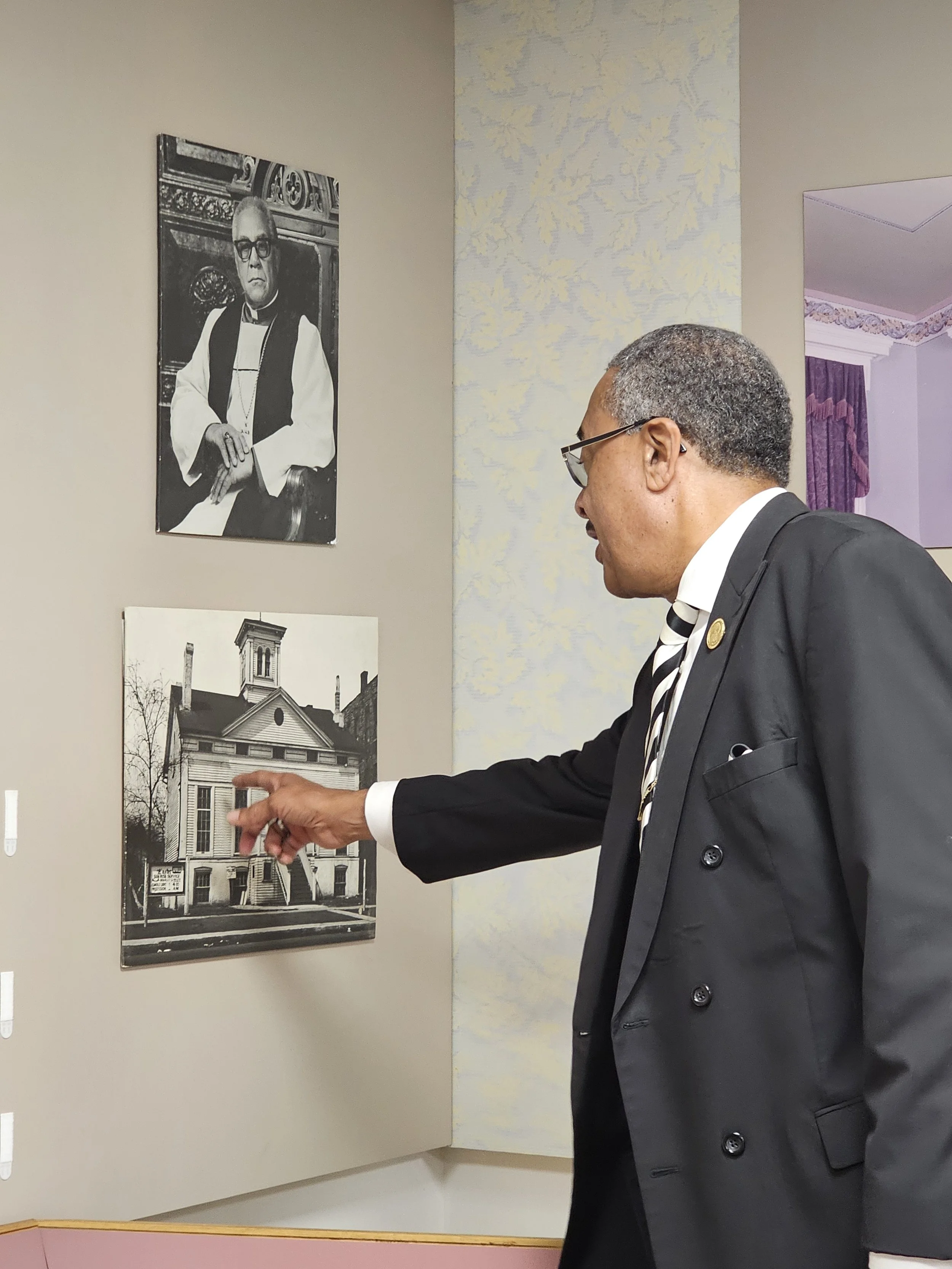 A man in a black suit and glasses points at a black and white photograph of a historic building displayed on a wall in an art gallery or museum.
