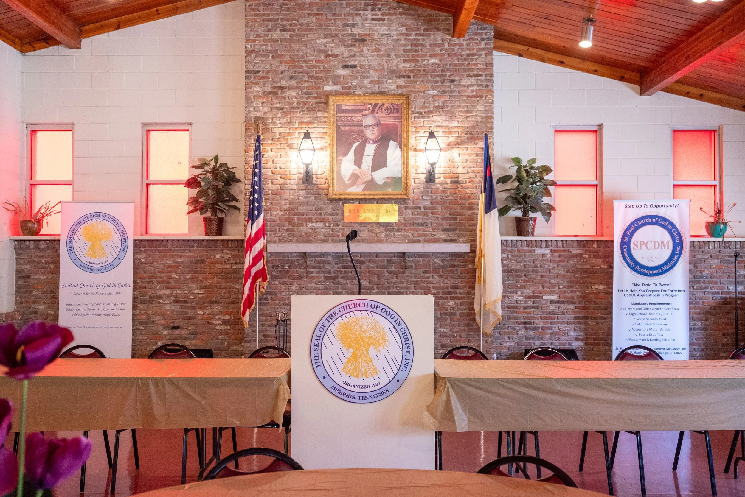 Interior of a church with brick and white walls, wooden ceiling beams, and a portrait of a man in clergy robes center on the wall, flanked by two American flags. Banners and potted plants are also visible.