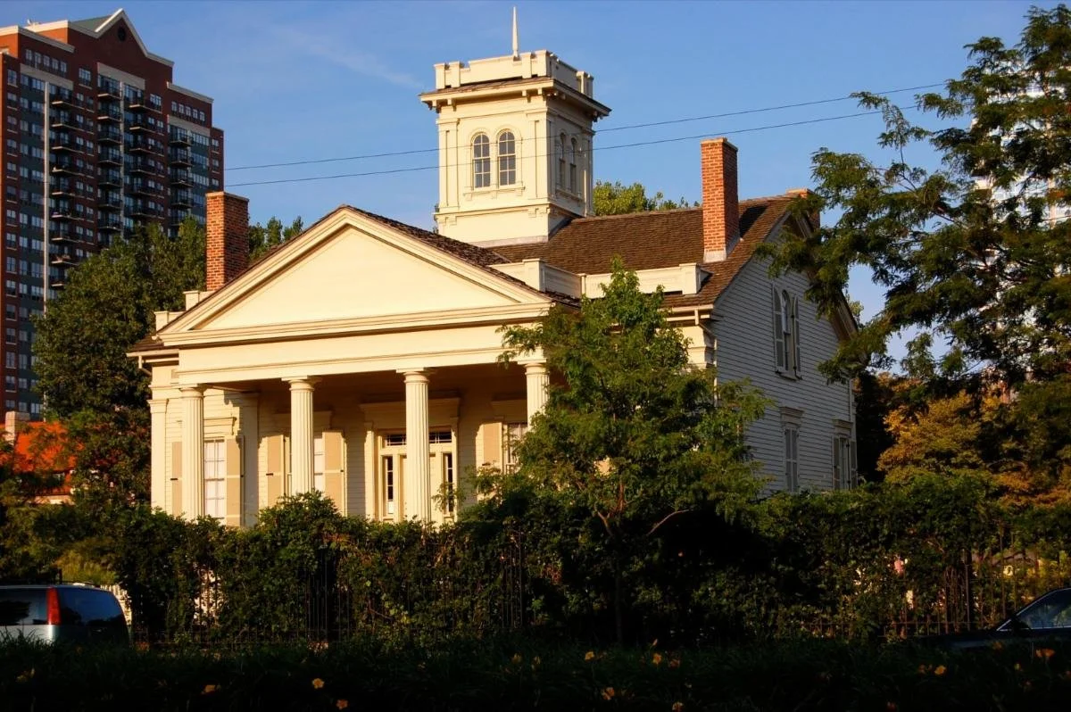 Historic white house with front columns, triangular pediment, and tower, surrounded by trees and bushes, with modern high-rise buildings in the background.