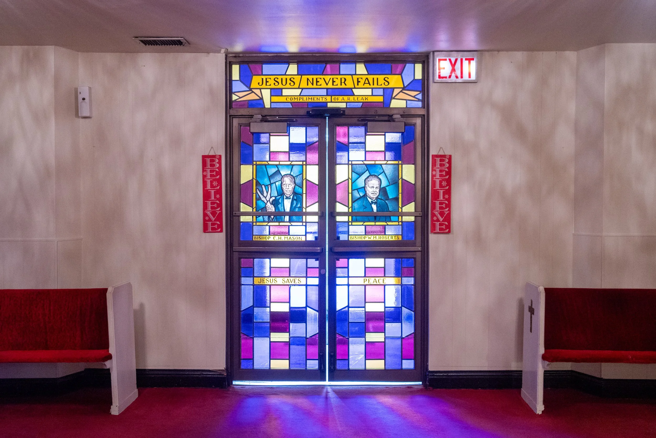 Stained glass church door featuring illustrations of two men with the words "Jesus Never Fails," "Jesus Saves," and "Peace" above and below, with red banners stating "Be Brave" on both sides.