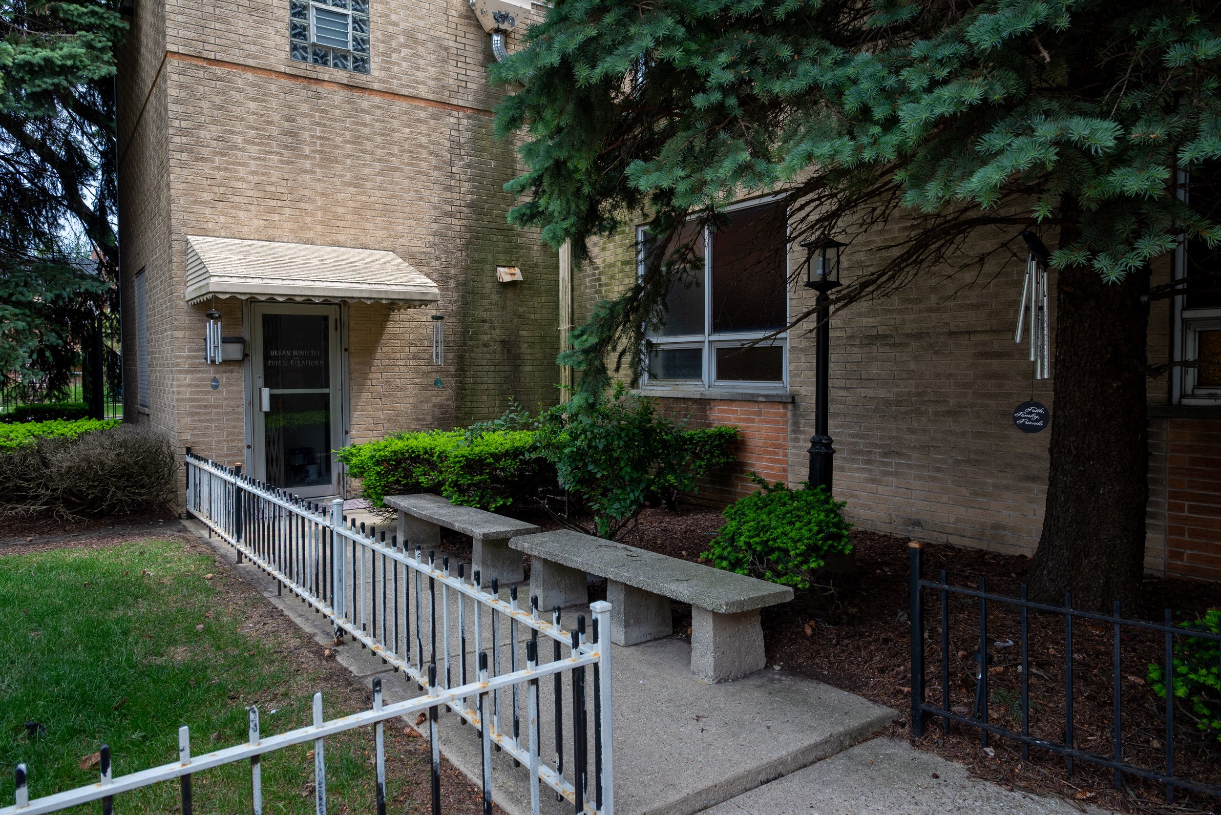 Apartment building with a brick exterior, green shrubs, and trees in front, featuring a black fence and metal wind chimes hanging from a tree.