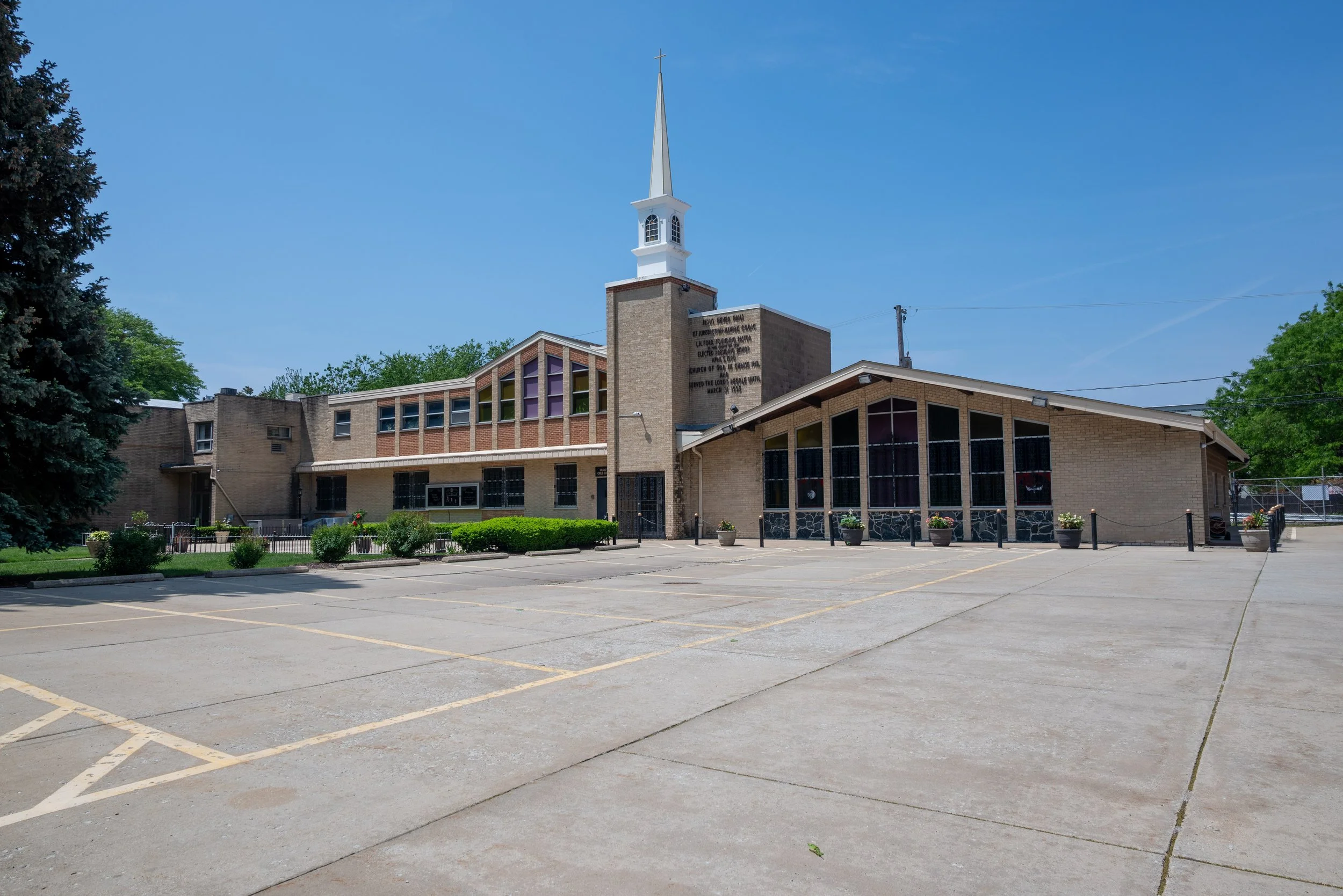 The image shows a church building with a tall white steeple, surrounded by trees and a mostly empty parking lot under a clear blue sky.