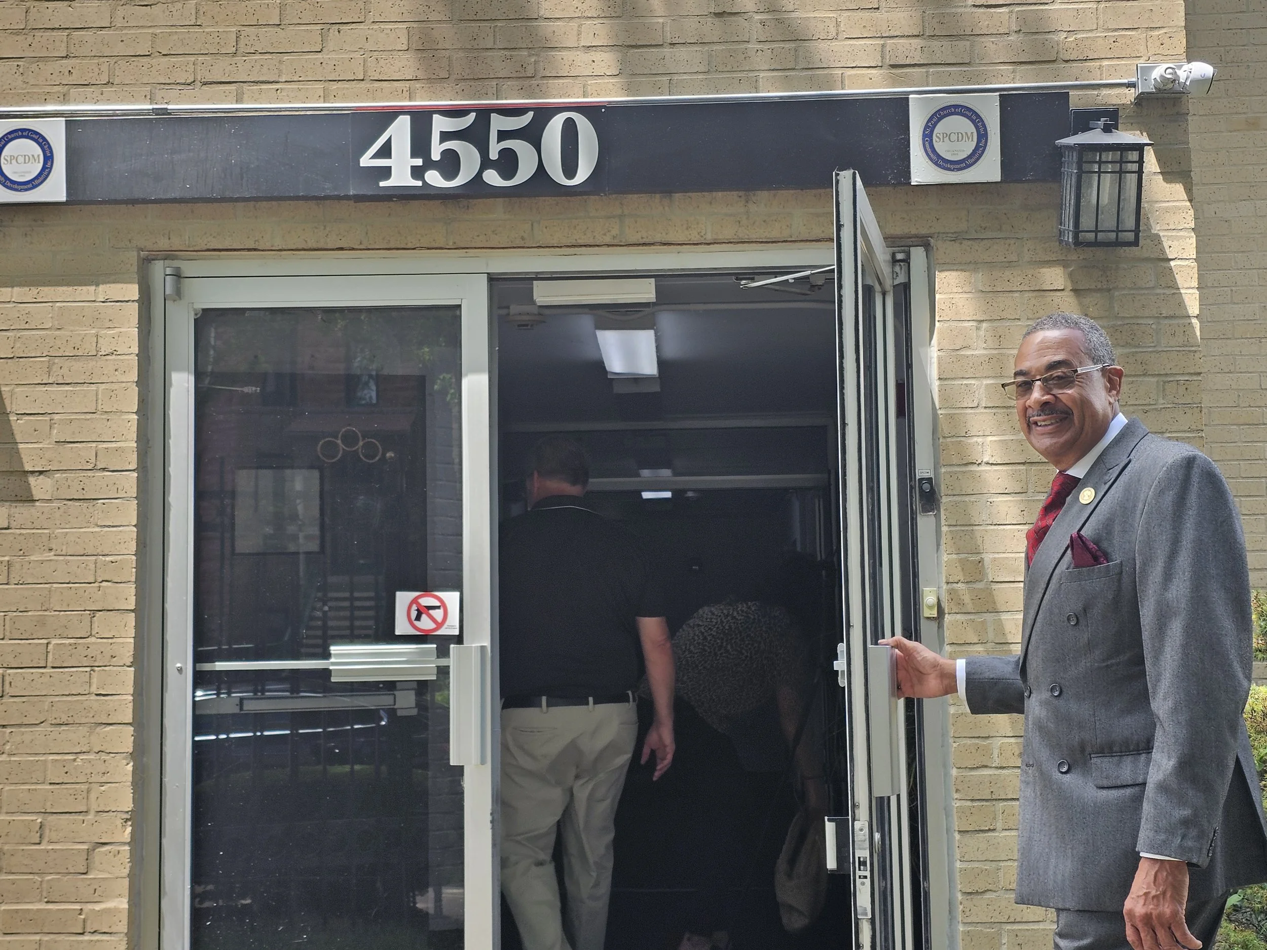 Man in a suit standing outside a building with the street number 4550 above the door, people entering through the door, and a no smoking sign on the door.