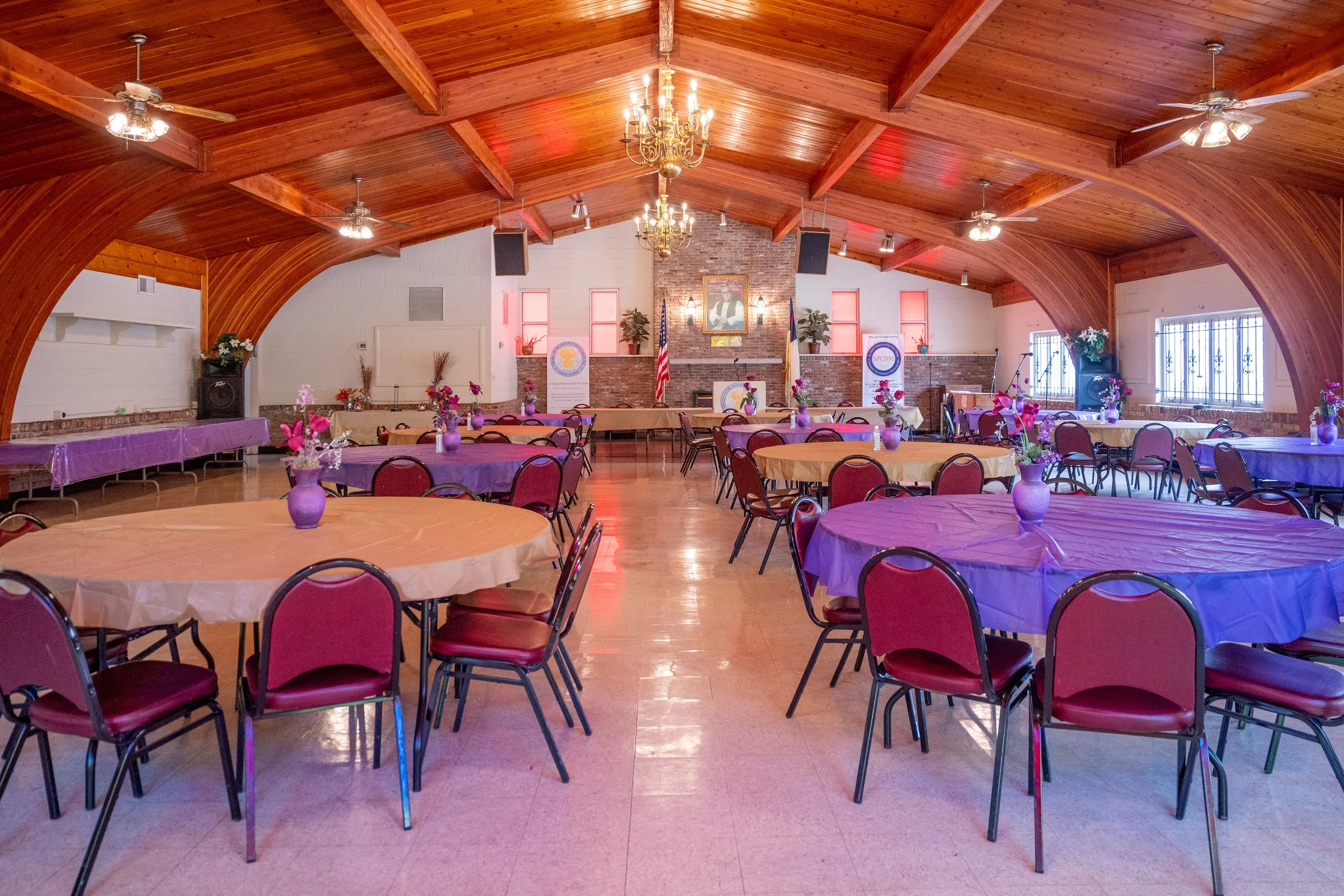 A large banquet hall with a high, curved wooden ceiling, chandeliers, ceiling fans, and a brick fireplace with framed portrait above. The room has round tables with purple and beige tablecloths, decorated with purple vases holding pink flowers. There