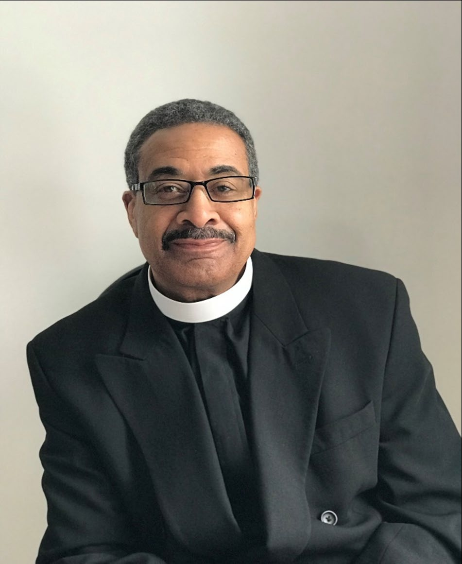 A man dressed as a priest, wearing a black suit with a clerical collar, sitting against a plain light-colored wall.