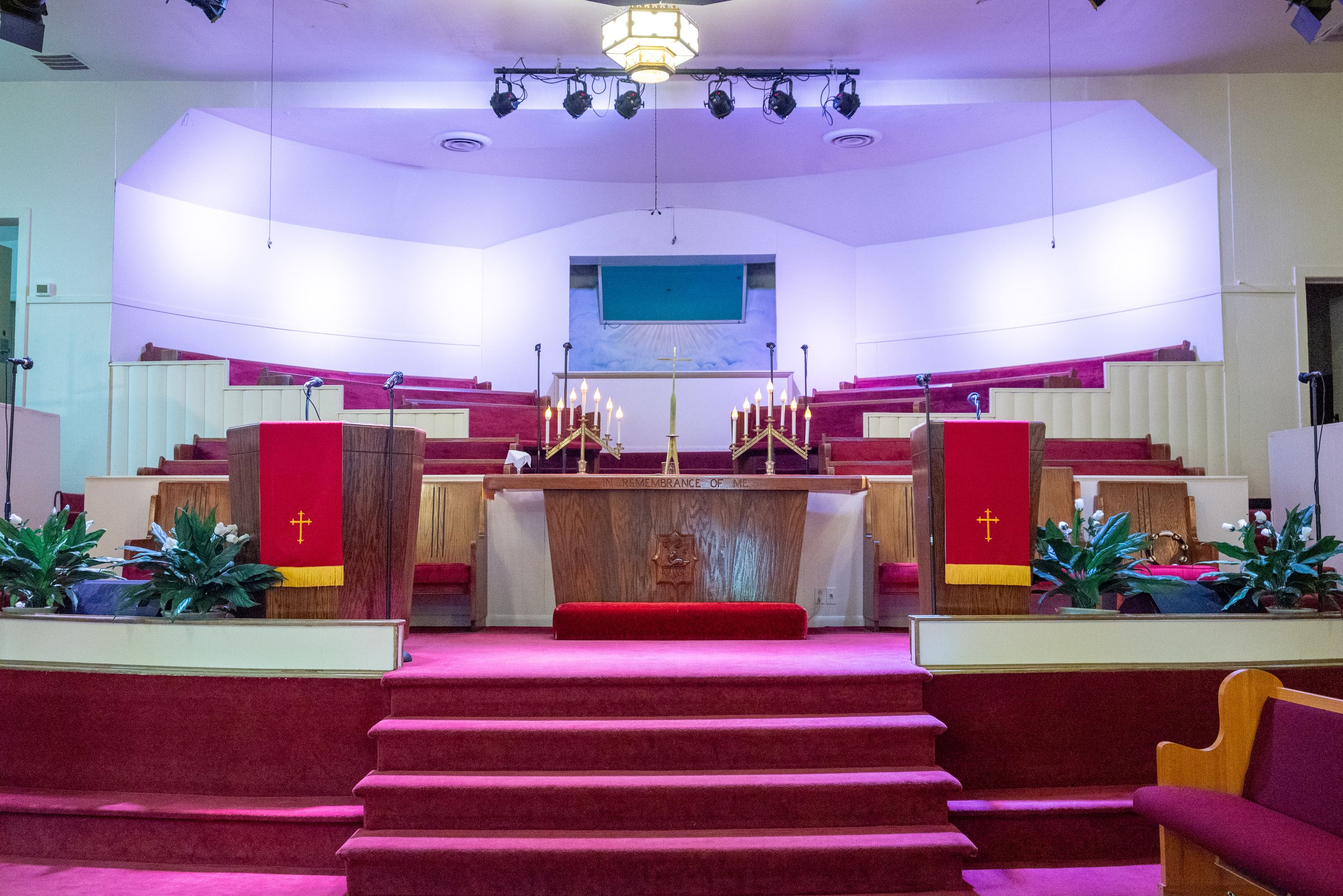 Inside view of a church altar with red carpeting, wooden stairs, and pews. The altar has candles, a cross, and plants. There are microphones on stands, a large screen in the background, and stage lighting equipment.