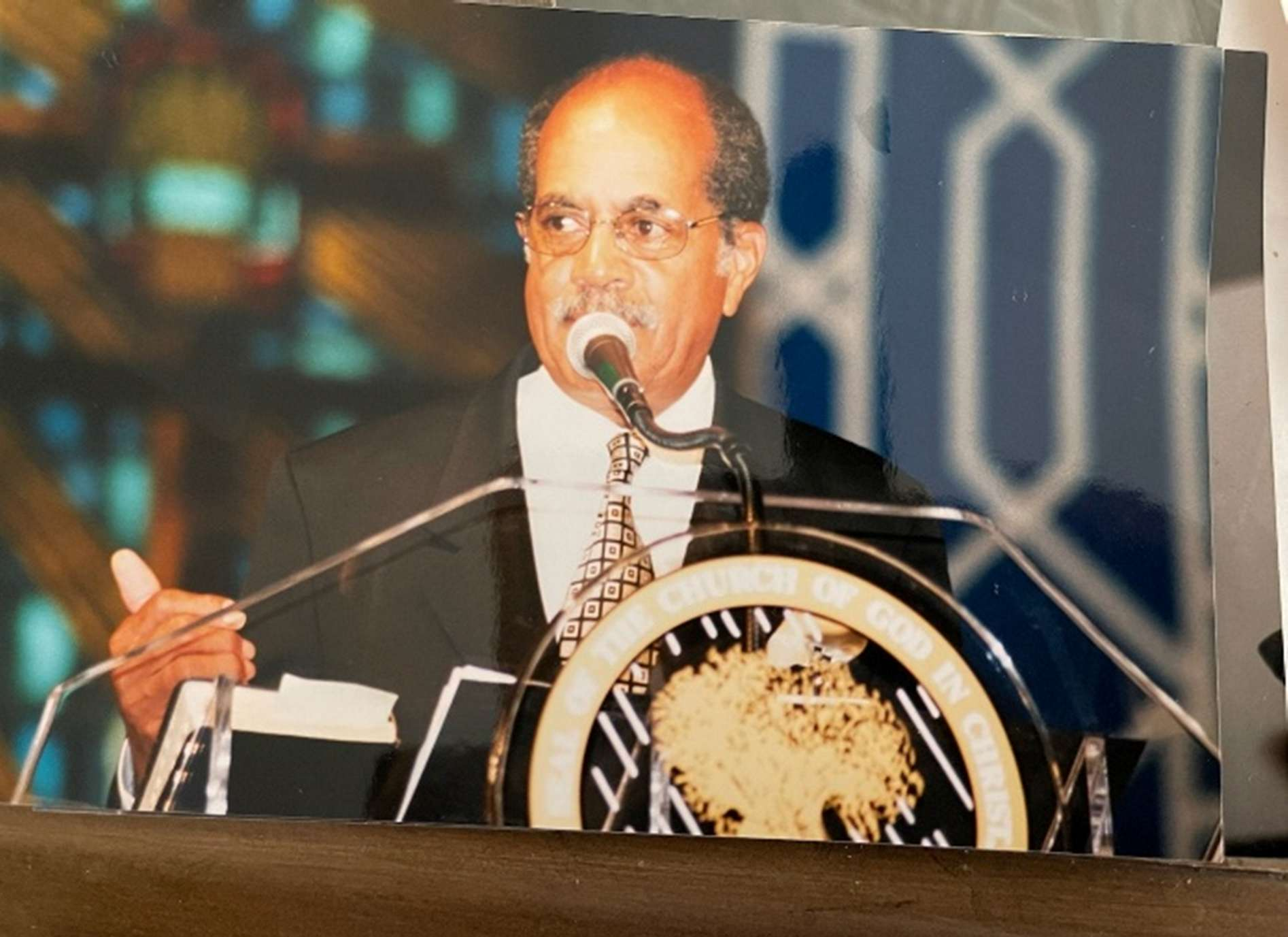A man in a tuxedo and glasses speaking at a podium during a formal event. There is a seal on the front of the podium and a background with patterned fabric.