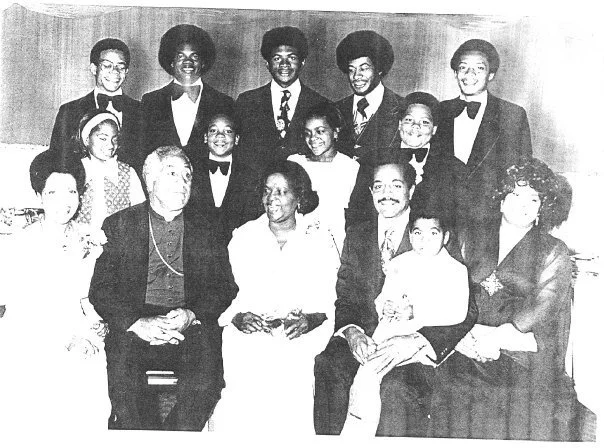 Black and white photo of a diverse group of people, both men and women, dressed in formal attire, posing in a studio setting.