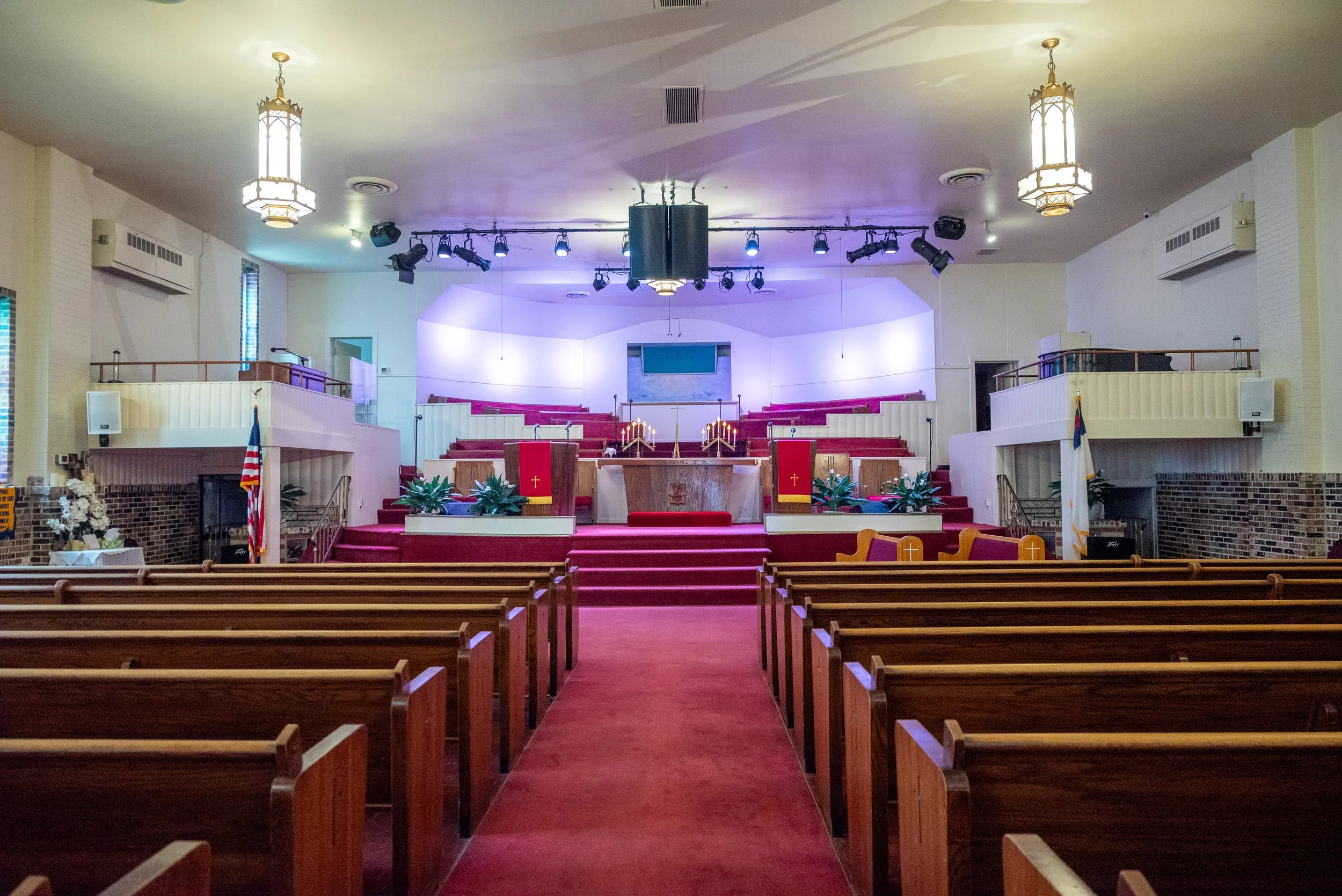 Interior of a church sanctuary with wooden pews, red carpeted aisle, stage with red and gold altar cloths, candles, and plants, stage lighting, chandeliers on the ceiling, and flags on either side.